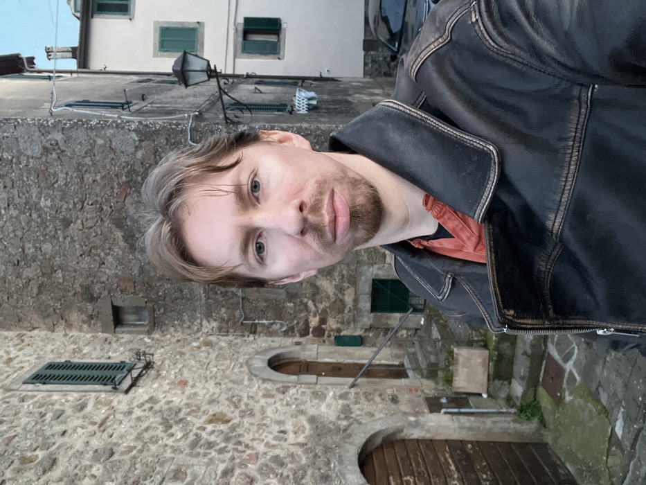An adult Caucasian male with light brown hair and a short beard, wearing a dark brown leather jacket over an orange-red shirt, is positioned centrally in the foreground. He looks directly at the camera with a neutral expression. The scene is an outdoor urban environment identified as Castel del Piano, Italy.
The background features historic architecture. To the immediate left, a multi-story building constructed of irregular light brown stone blocks displays two arched wooden doorways, a set of stone steps, and a green wall-mounted mailbox. Above these, a window with dark green louvered shutters is visible, along with a smaller, recessed rectangular opening. To the right and further back, a narrower street or alleyway leads past a lighter-colored building (possibly stucco) with multiple windows, each featuring dark green louvered shutters. An ornate black metal street lamp extends from this building's wall. A dark-colored modern car is partially visible parked on the street beyond the lamp post. The ground is paved with uneven cobblestones or flagstones, with patches of green moss visible in crevices. The sky above is light and clear.