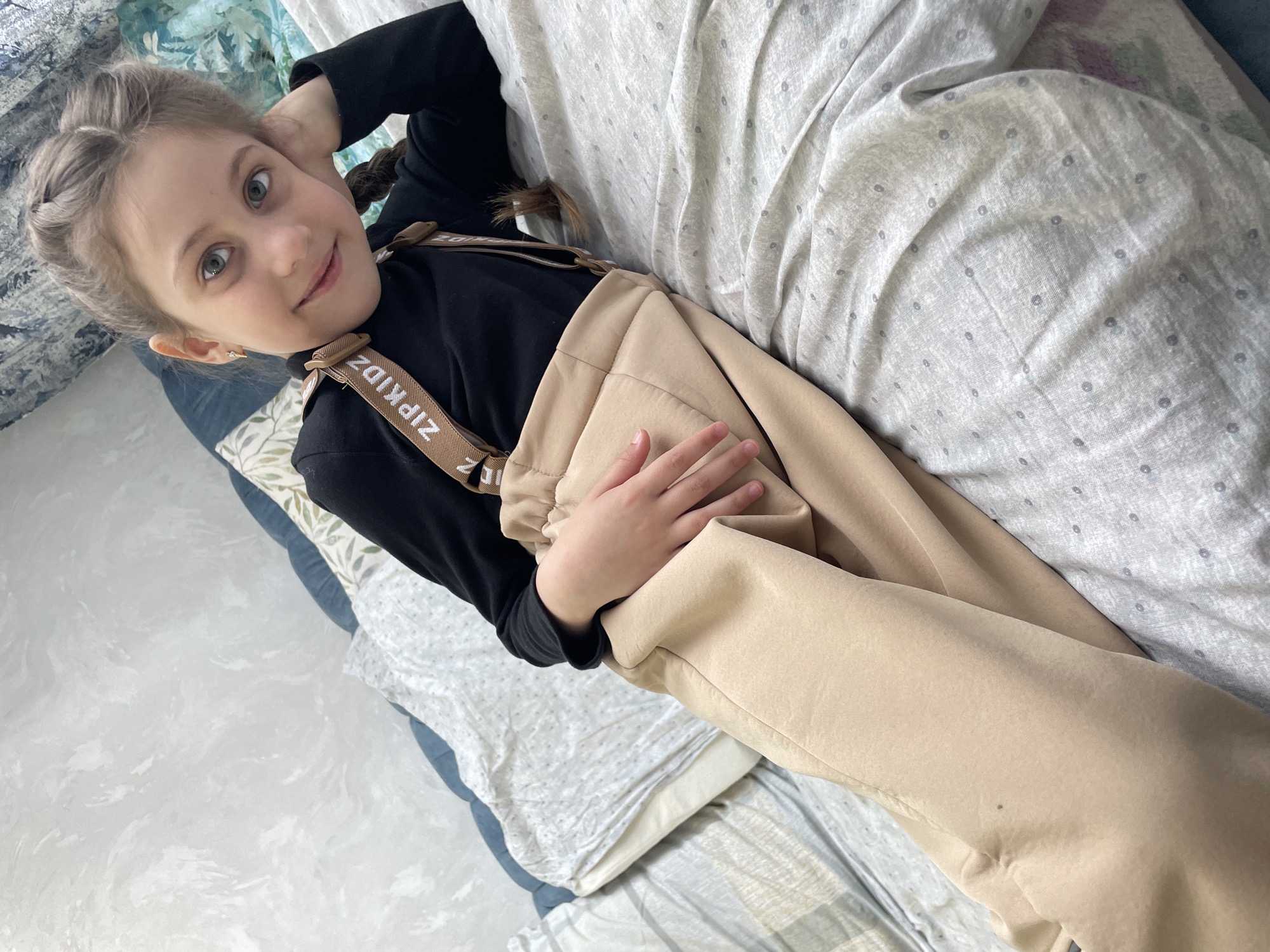 A young girl lies on a bed covered with patterned bedding, possibly in a residential setting. She wears a black long-sleeved shirt with beige overalls labeled "ZIPKIDZ". Her hair is styled in braids. The scene is indoors, with diffused natural light. The location is unspecified, with no distinct markers indicating it is Izhevsk, Russia.