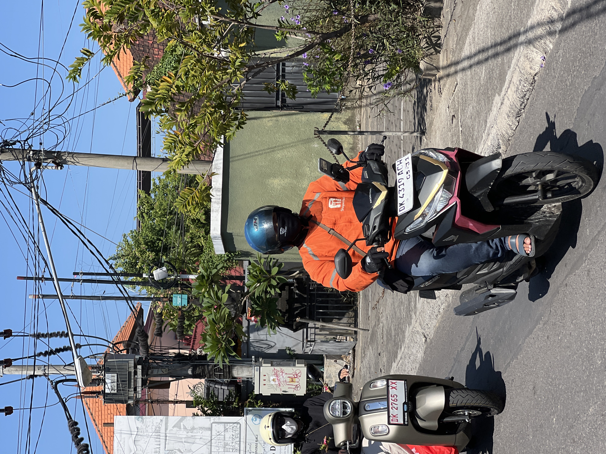 Two individuals are riding motorcycles on a street in Denpasar, Indonesia. The motorcycle on the left is a light brown Yamaha scooter with a license plate reading "DK 2765 XX". The rider is wearing a black long-sleeved shirt, white helmet, and a white face mask. The motorcycle on the right is a dark-colored Honda with a license plate reading "DK 4319 ACH". The rider is wearing an orange long-sleeved shirt with a Shopee Food logo, blue jeans, a blue helmet, and a black face mask. Both riders are wearing gloves. The street appears to be paved with asphalt and has a concrete curb. In the background, there are buildings with orange tiled roofs, lush green trees, and numerous electrical wires strung across utility poles. The sky is clear and blue, indicating a sunny day. A transformer box is visible on a utility pole. The scene captures a typical urban environment in Denpasar, likely during daytime hours.