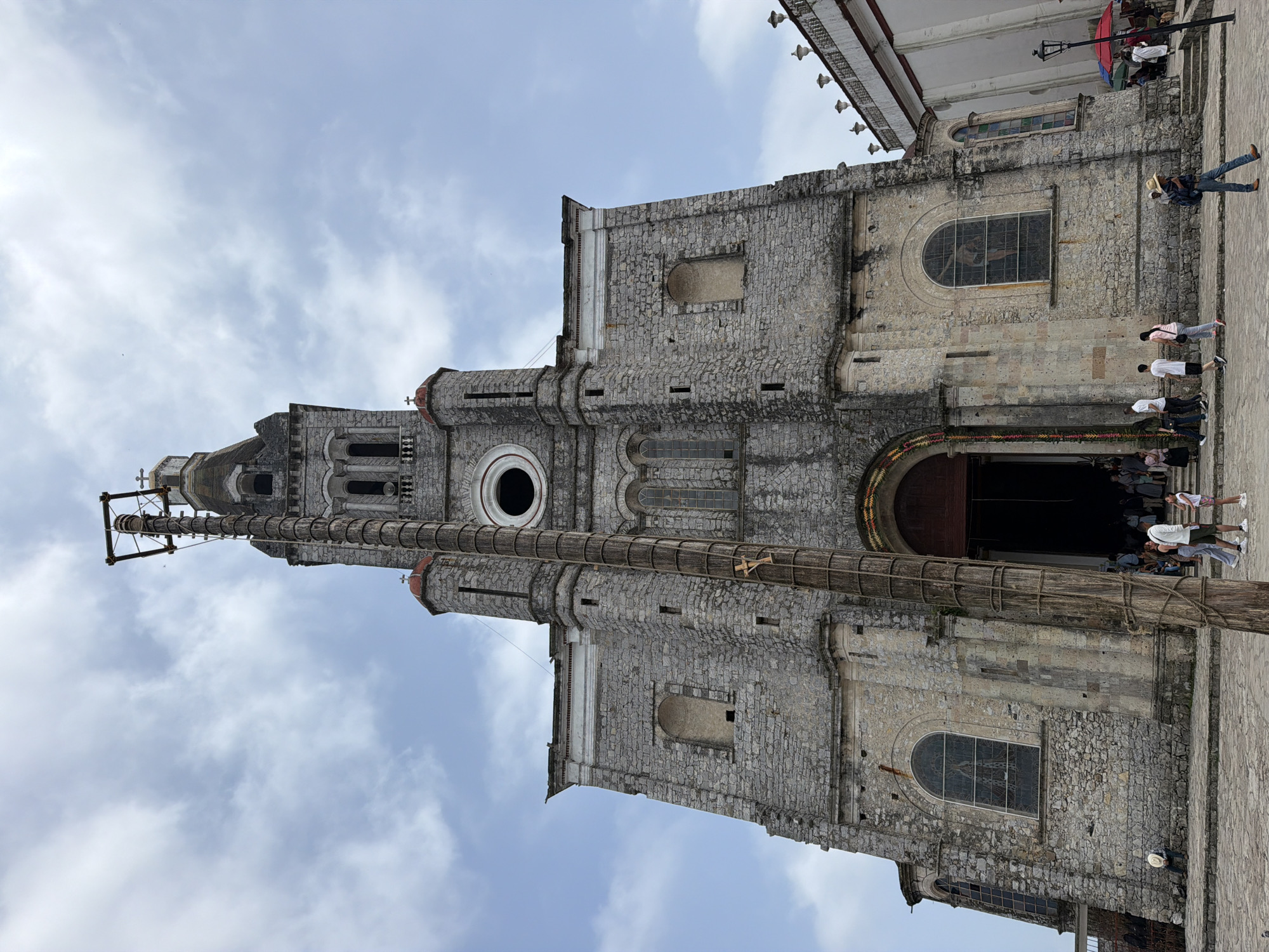 The image shows the Iglesia de San Francisco de Asís (Church of San Francisco of Assisi) in Cuetzalan, Mexico. The church is constructed of gray stone with a tall tower. A tall wooden pole stands in front of the church. People are standing and walking near the church entrance. The sky is cloudy.