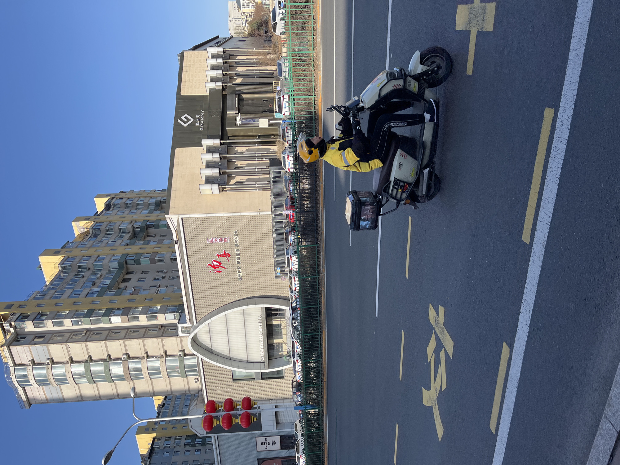 The image captures a street scene in Changchun City, China. A person in a yellow jacket and helmet is seated on a white scooter with a delivery box on the back, stopped on the road. The street has white and yellow lane markings, and Chinese characters painted on the road surface indicate a pedestrian crossing. Behind the scooter, a green fence separates the road from a row of parked cars and the sidewalk. Buildings flank the street. One building displays the name "GRAMMY" in English, along with Chinese characters. Another building is topped by the Chinese characters for a hot springs and spa resort. Traffic lights with red lanterns are also visible. The sky is clear blue.