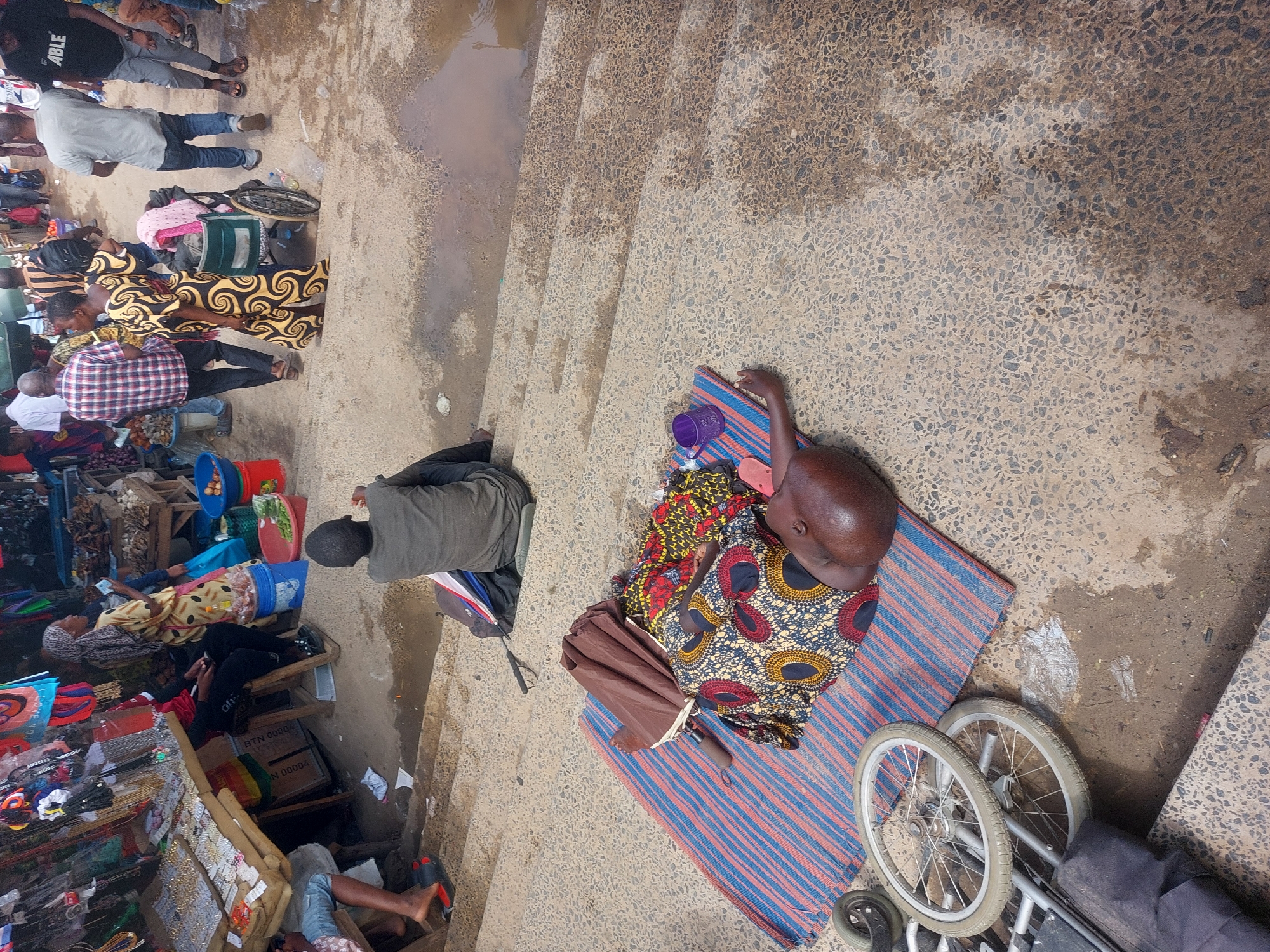 This image captures a high-angle view of a busy outdoor market scene in Dar es-Salaam, Tanzania. In the foreground, towards the right side of the frame, a person with dark skin is lying on their left side on a blue and red striped mat. They are dressed in a vibrant, patterned traditional African fabric. Next to them on the mat are a purple plastic cup and a folded brown umbrella. Two metal wheels, possibly belonging to a wheelchair or handcart, are visible near the mat. Further up and to the left, another person is seated on the ground, clad in dark attire.
The left side of the image is densely populated with numerous individuals, predominantly adults, standing and moving amidst various market stalls. These stalls display a variety of goods, including colorful plastic buckets, jewelry, and what appears to be produce. People are seen wearing diverse clothing, from plaid shirts and jeans to brightly patterned traditional dresses.
The activity depicted is that of a bustling street market, with individuals engaged in walking, selling, and possibly buying. The person on the mat appears to be resting or perhaps awaiting assistance. The ground is a textured, light-colored aggregate surface, marked by several dark, damp patches and puddles of water, suggesting either recent rain or spillage. The bright, natural lighting indicates it is daytime.
Visible text includes the word "ABLE" partially printed on a black t-shirt worn by a person in the upper left of the frame. On a wooden crate in the lower left, "BTN 00004" and "N 00004" can be seen. The overall scene conveys the vibrant and dynamic atmosphere of a local market.