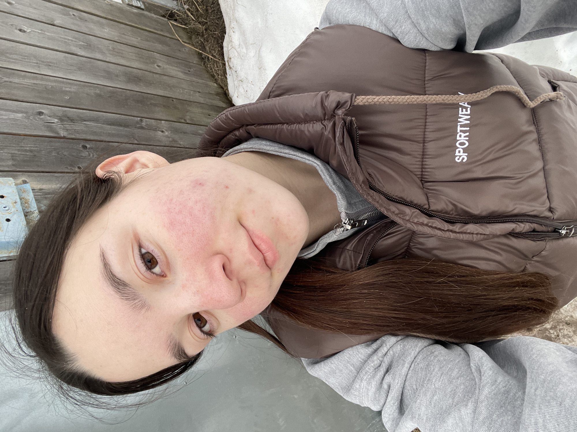 A young woman takes a selfie outdoors. She has long brown hair, brown eyes, and some visible acne on her left cheek. She is wearing a grey hooded sweatshirt and a brown puffy vest with the word "SPORTWEAR" printed on it. The background includes wooden planks, some snow, and a light grey, possibly icy surface below. The location is potentially Ольховка, Russia.