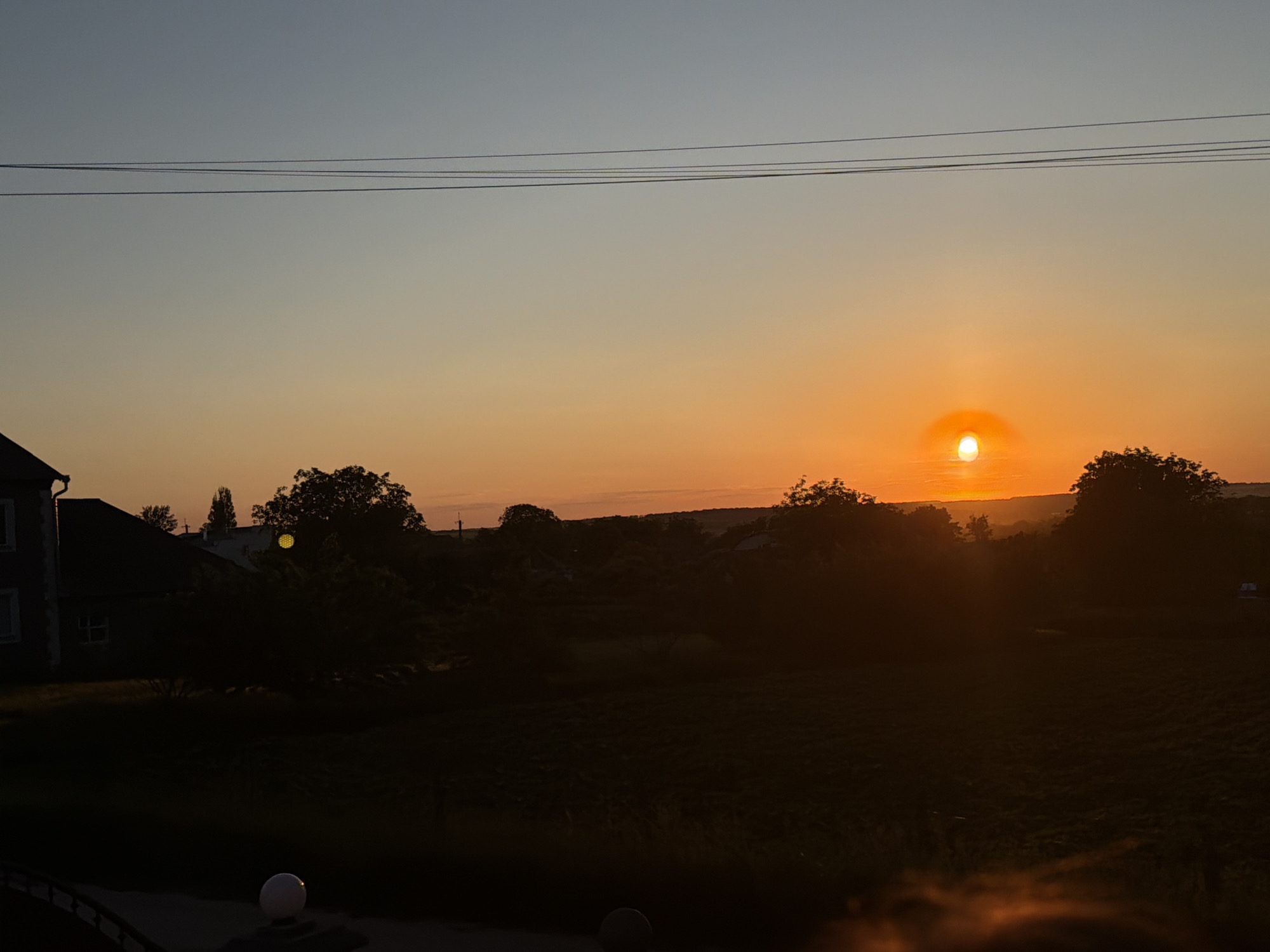 The image depicts a sunset over a rural landscape. In the foreground, a dark silhouette of a building or house is visible on the left, with a portion of a fence and a spherical light fixture. The middle ground features a silhouetted tree line and the dark shapes of more buildings and trees. A field of dark vegetation occupies the lower portion of the image. The sky is a gradient of soft blue at the top, transitioning to a vibrant orange and yellow as it approaches the horizon, where the sun is setting. The sun is a bright, glowing orb with a halo effect. A few power lines are visible in the upper sky. The scene appears to be set in a village or rural area.