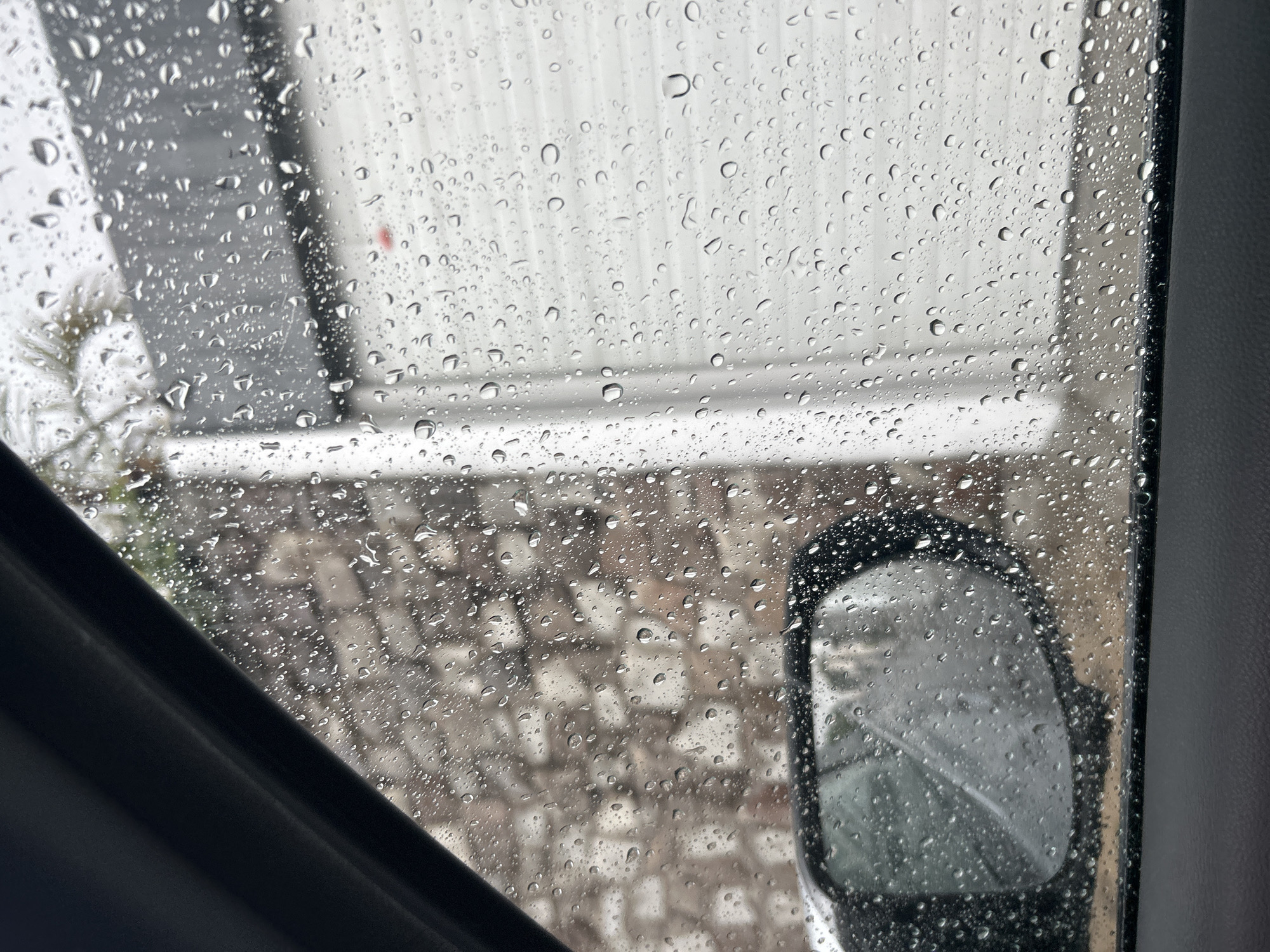 The image shows a view from inside a vehicle on a rainy day. The rain is visible on the car window, and the outside scene is blurred due to the rain. Outside the window, there is a building with a corrugated metal facade and a lower section of brick or stone. A side mirror of the vehicle is visible in the lower right corner of the frame. The location appears to be an urban or suburban area.