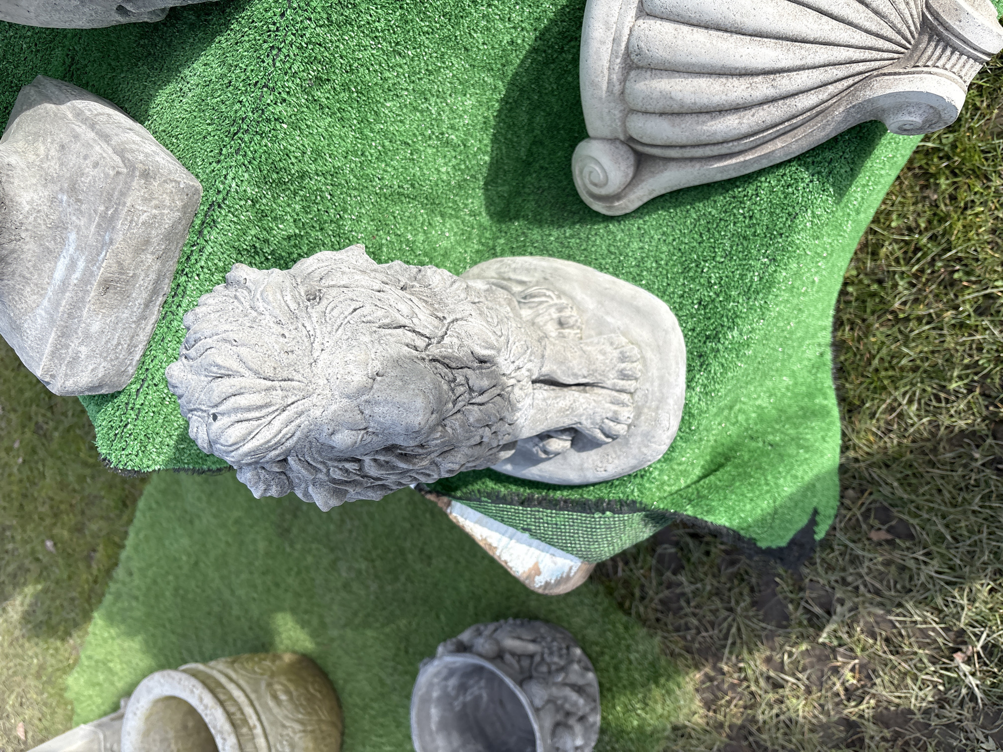The image depicts a collection of stone garden ornaments displayed on artificial green turf. Dominating the scene is a lion statue, viewed from above, its head and paws clearly visible. Adjacent to the lion are other stone elements, including architectural fragments and a vase or planter. The setting appears to be outdoors, with natural grass and soil visible around the artificial turf, suggesting a garden or display area. The image's origin in Chirk, United Kingdom, implies this could be a garden center or a residential garden in that locale.