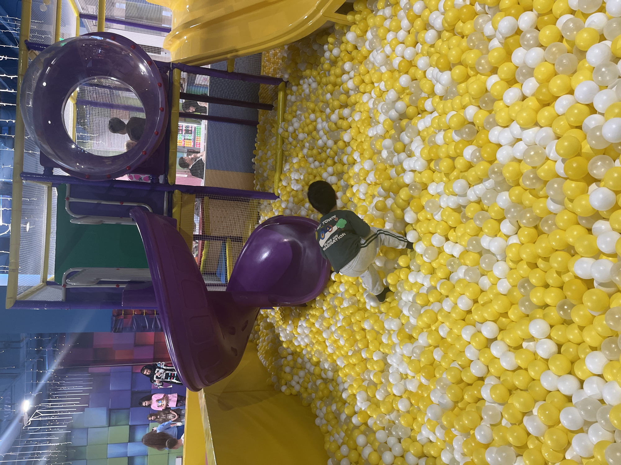 A child is standing in a large ball pit filled with yellow and white balls. The pit is adjacent to a yellow platform and a purple slide. The background includes play structures with netting, and a group of people can be seen in the lower left corner. The location may be in Sochi, Russia.