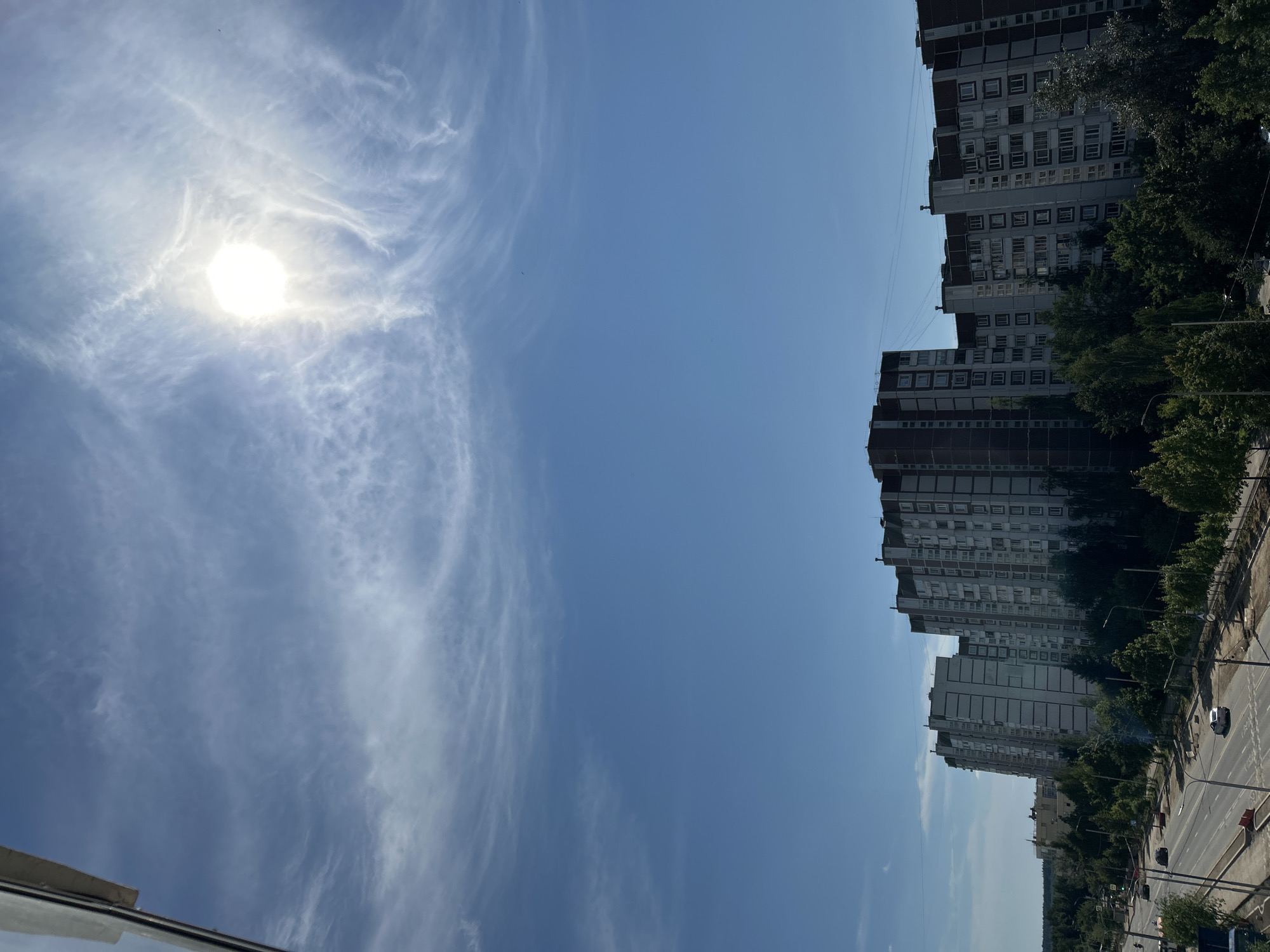 This is a vertical, eye-level, outdoor, daytime, full shot of apartment buildings in Zelenograd, Russia. The sun is visible in the upper right corner of the frame, partially obscured by wispy clouds. In the foreground, a multi-lane road runs horizontally across the bottom of the image, with several cars visible. A row of trees lines the road, separating it from a series of tall, multi-story apartment buildings that dominate the mid-ground and background. The architecture of the buildings appears to be typical of Soviet-era mass housing. The sky is a clear blue, with a few scattered clouds. The overall impression is of a suburban or residential area.