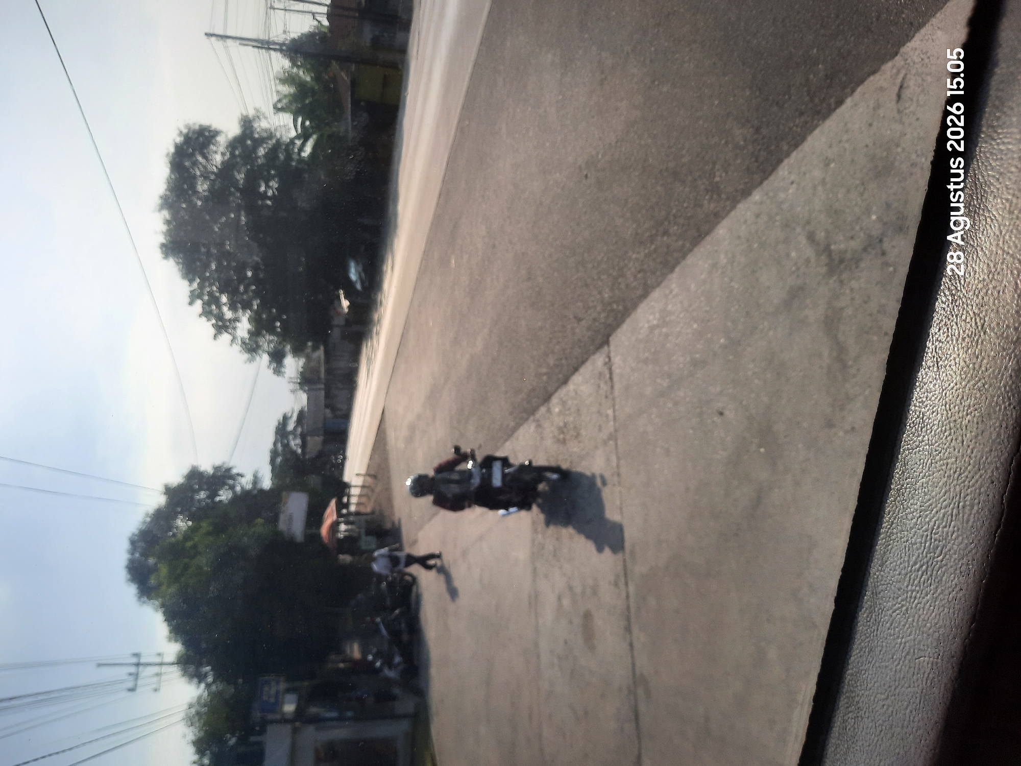 The image depicts a street scene in Gending, Indonesia, on August 28, 2026, at 3:05 PM. A person on a motorcycle, wearing a helmet and a backpack, is riding on the right side of a paved road. Another person is walking on a sidewalk on the left side of the road. There are parked motorcycles, trees, and buildings visible in the background. The sky is clear and blue. The image appears to have been taken from a vehicle.