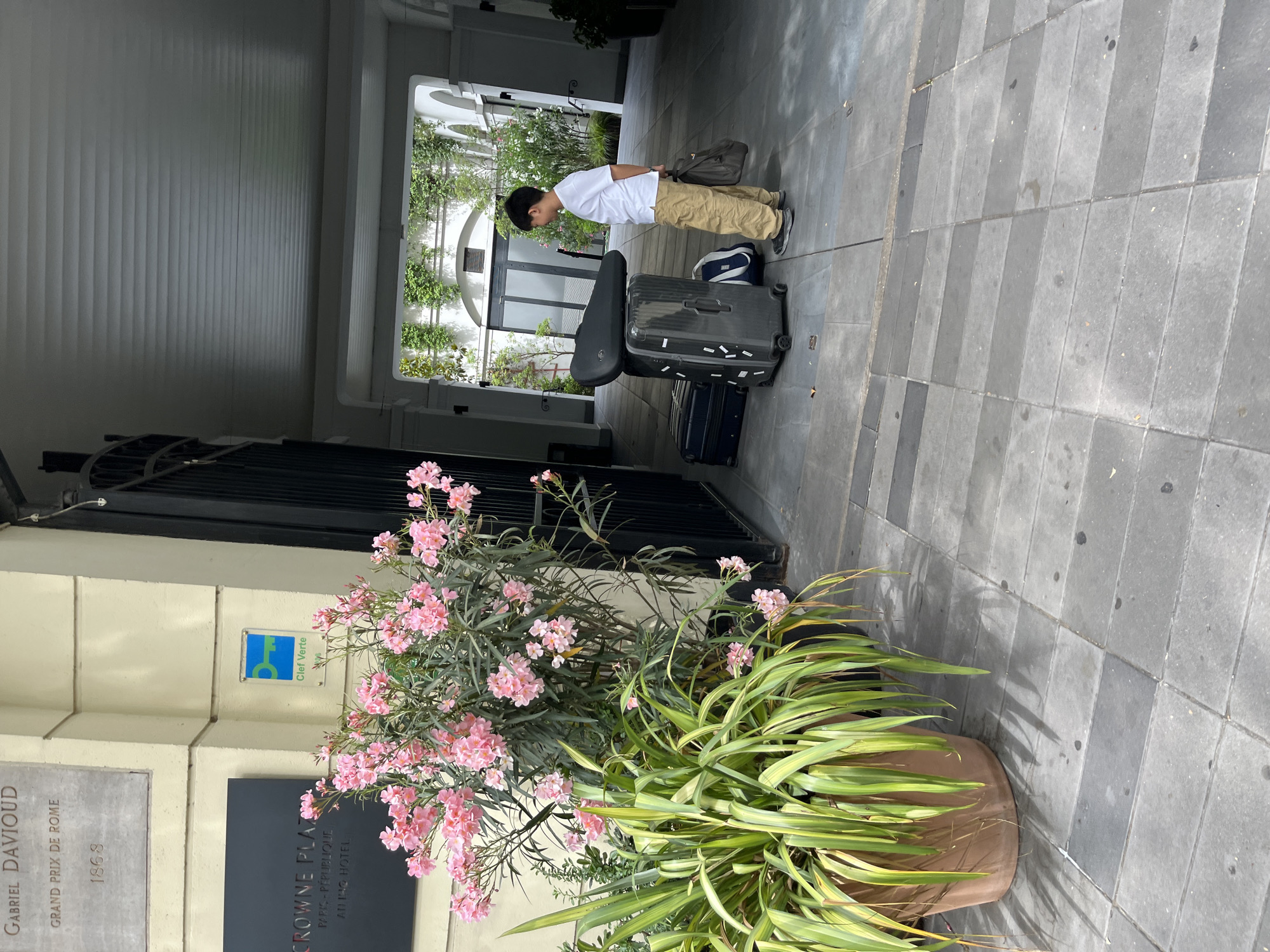 The image shows a street scene in Paris, France, outside the Crowne Plaza Paris – République hotel. A young boy stands next to a stack of luggage, including a large black suitcase, a dark blue suitcase, and a black bag on top of the suitcase. The boy is wearing a white t-shirt and tan pants, and he is holding a dark brown bag in his right hand. In the foreground, there is a large potted plant with pink flowers and long green leaves. Behind the boy and luggage, a dark metal gate is visible, and further back, an archway leads into a courtyard with greenery. A sign for "Clef Verte" (Green Key) is mounted on the building to the left of the gate. A plaque above that reads "GABRIEL DAVIOUD GRAND PRIX DE ROME 1868" is also visible. The ground is paved with gray tiles.
