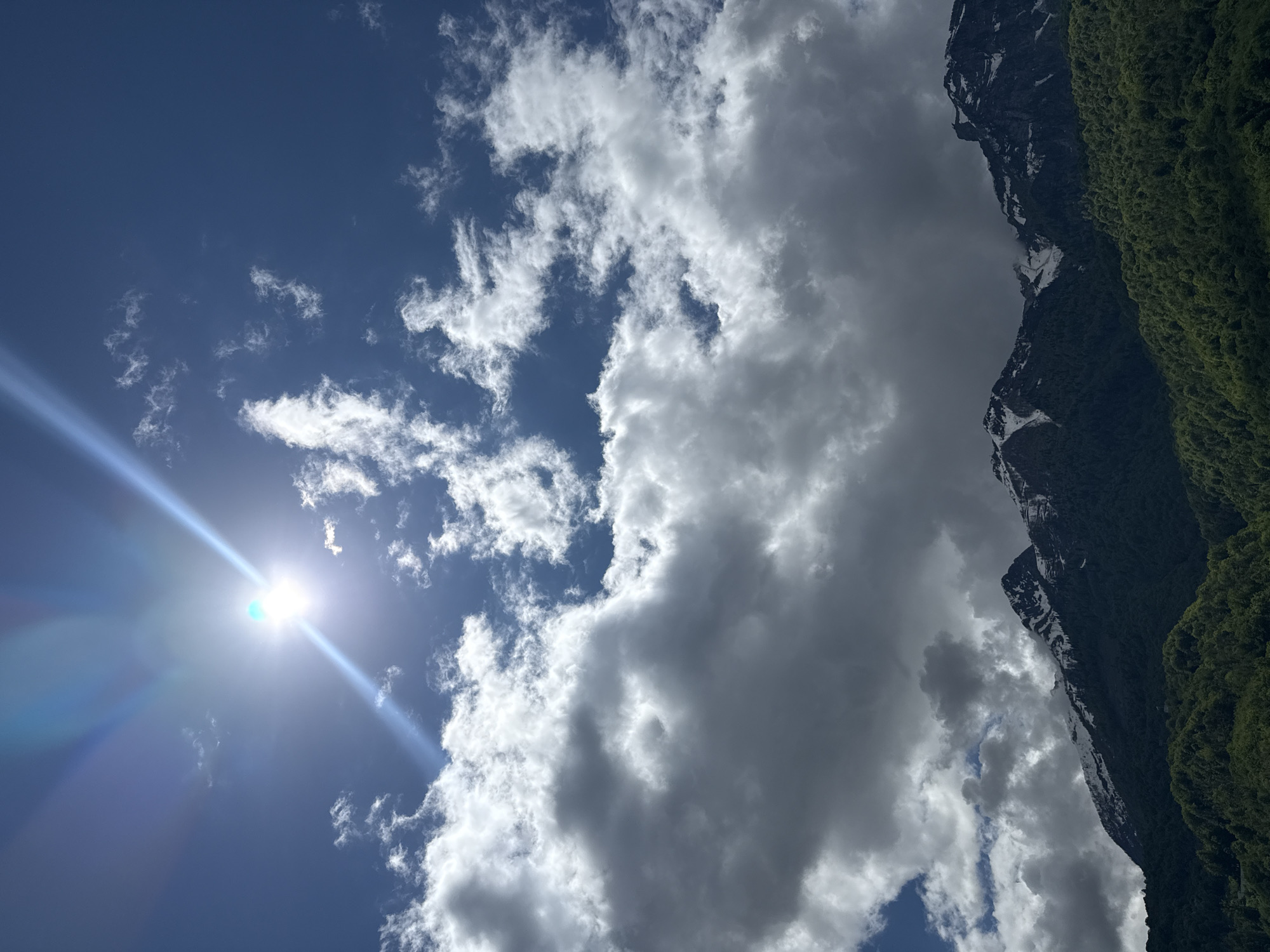 The image captures a view of a sky partially obscured by clouds and a portion of a mountain covered with greenery. The sky is a deep blue, with bright sunlight visible in the lower left corner. Several white, puffy clouds dot the sky, while a large, grey cloud formation dominates the right side of the image, partially obscuring a tall, dark mountain range with some snow patches. Lush, green vegetation covers the lower part of the mountain. The scene is likely set in a mountainous region, potentially Krasnaya Polyana, Russia, given its topography, which is known for having similar natural features.