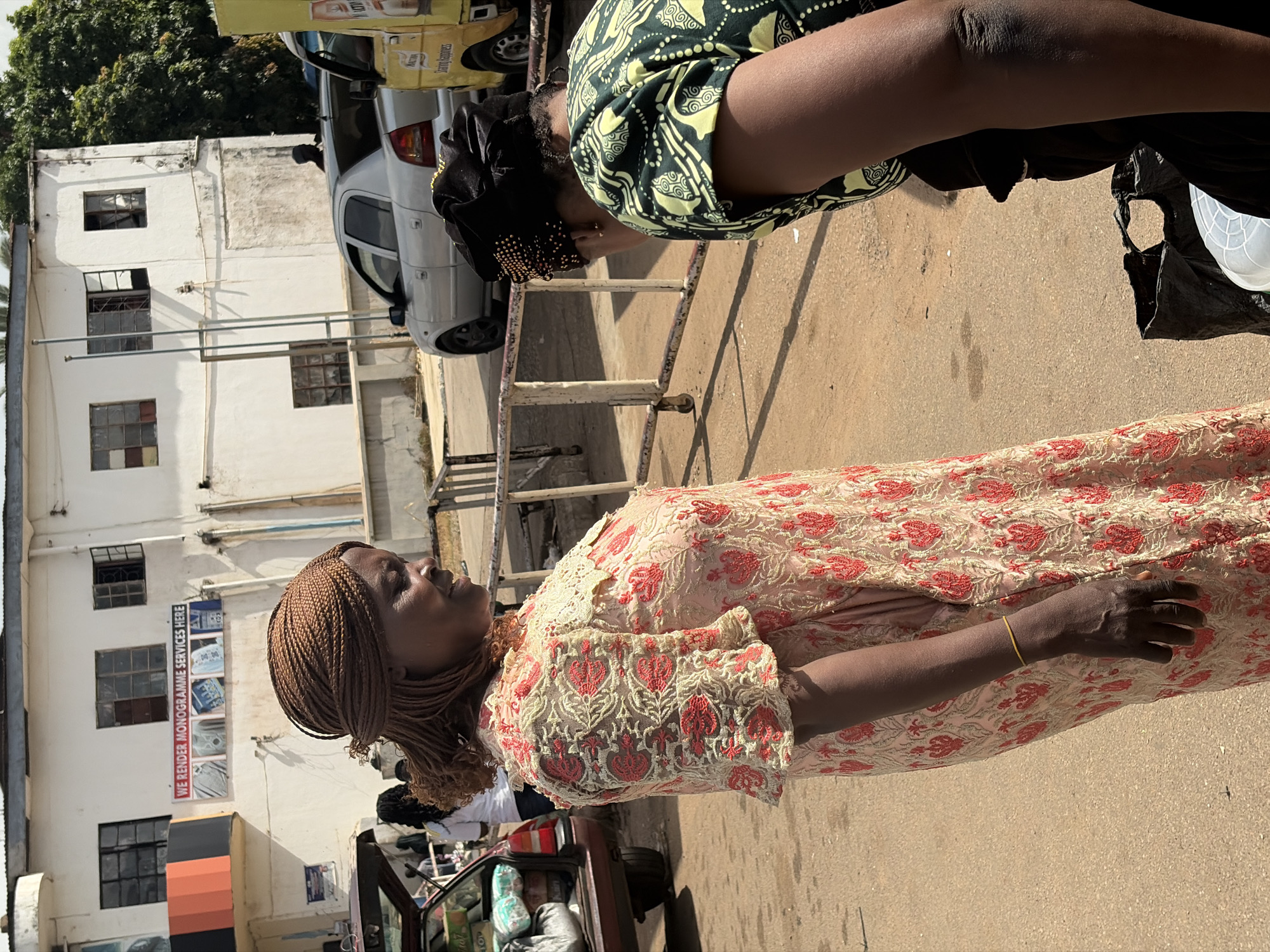 The image depicts an outdoor scene on a street in Jos, Nigeria, likely in a commercial area. In the foreground, two women are visible. The woman on the left is facing towards the right and is wearing a light-colored dress with intricate red floral embroidery. She has braided brown hair pulled back. The woman on the right is seen from behind, facing towards the left, and is wearing a dark headscarf adorned with gold beads and a green and white patterned garment. She is bending slightly.
Behind the women, a light-colored sedan is parked, and further back, a yellow commercial vehicle is visible. A white building with multiple windows, some of which appear broken or boarded up, forms the backdrop. A sign on the building reads "WE RENDER MONOGRAMME SERVICES HERE" and displays images of embroidered items. A metal railing with wheels is positioned between the foreground and the vehicles. The ground is paved with a rough, sandy surface. The lighting suggests it is daytime, with distinct shadows. The overall impression is that of a busy street scene.