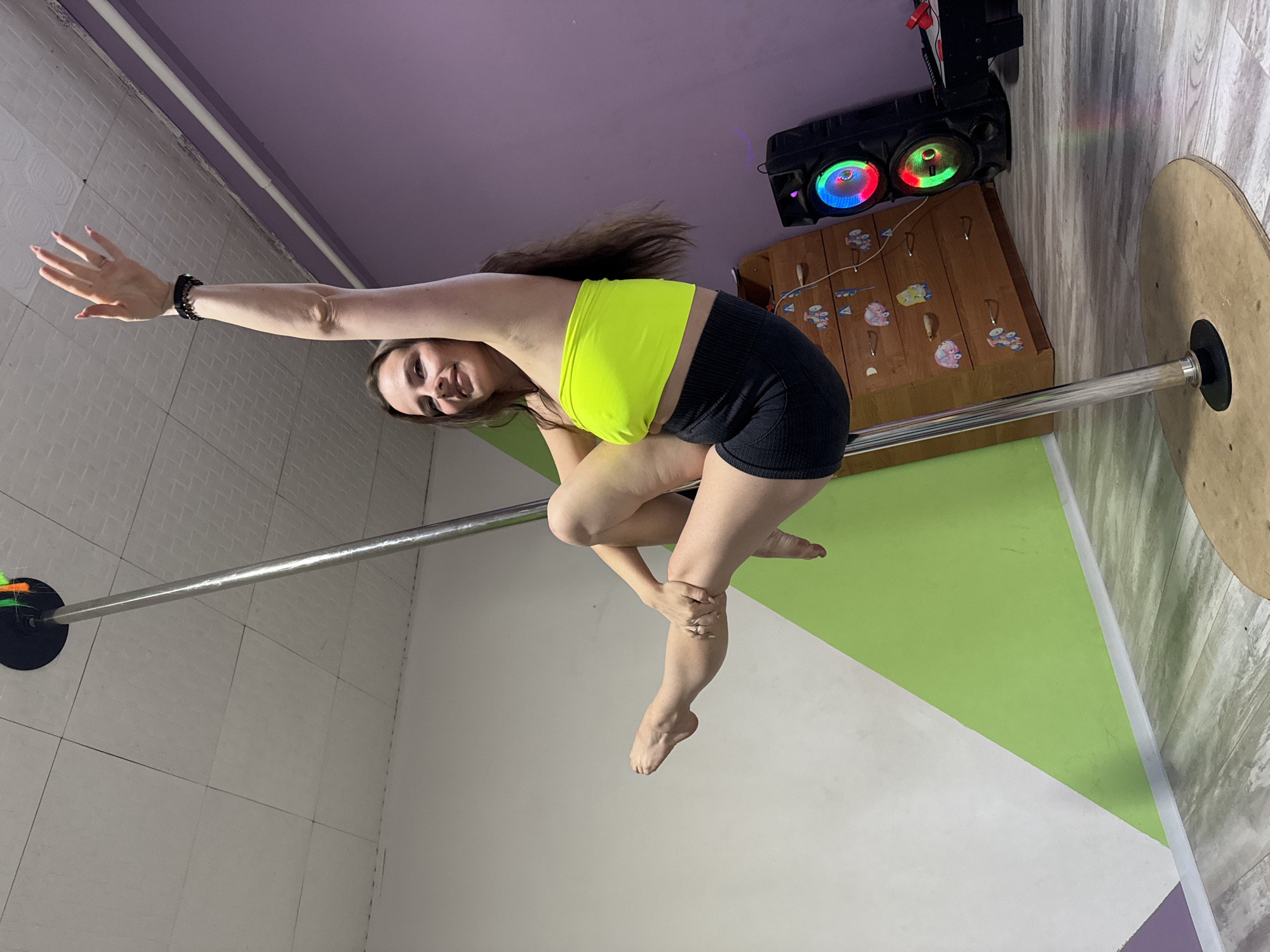 A young woman with long, dark hair is captured mid-pose on a metal pole. She is wearing a bright neon yellow crop top and black shorts. Her left arm is extended upwards, with her palm open, while her body is contorted in a dynamic pose. Her legs are bent and crossed, with her right hand holding her left knee. She has a slight smile on her face, and her hair is flying back, indicating movement.
The background suggests an indoor studio or recreational space. The walls are a pale grey and lime green, with a purple ceiling. A wooden cabinet with decorative knobs and possibly toys is visible to the right, along with a large black speaker system with colorful, pulsing lights. A wooden disc is attached to the base of the pole. The lighting in the room appears to be artificial, with a slight rainbow effect visible on the wall from the speaker lights. The image is oriented vertically, suggesting it was taken with the camera held in a portrait orientation. The overall impression is one of athleticism and performance.