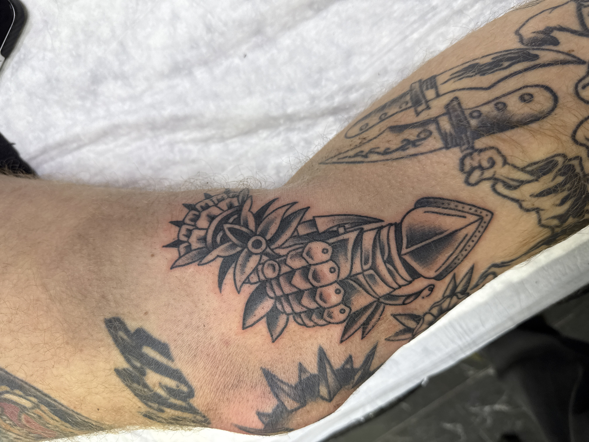 A close-up shot focuses on a person's light-toned, hairy arm, showcasing a freshly done tattoo on the inner forearm. The main tattoo features a stylized gauntlet (armored glove) holding a bouquet of flowers and berries. Rendered in black and grey, it displays bold outlines, solid black areas, and detailed shading, characteristic of American traditional or neo-traditional tattoo styles.
Around this central piece, other tattoos are visible, suggesting a sleeve. To the upper right, part of a dagger or knife tattoo can be seen, also in black and grey. Below the main tattoo, fragments of other blackwork designs are present. The skin immediately surrounding the new tattoo appears slightly red, indicating its recent application.
The arm is resting against or held near a white, textured fabric, possibly a towel or sheet. The setting appears to be indoors, likely in a tattoo studio or similar environment, given the focus on the fresh artwork. No specific time of day, weather, emotions, or location cues are discernible from the image, nor is there any visible text.