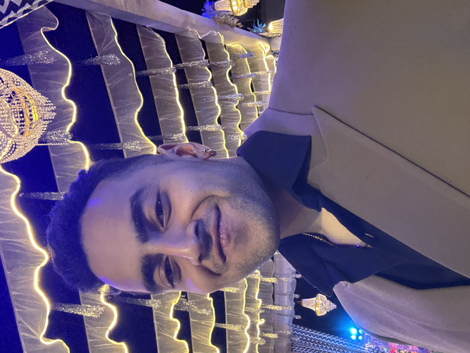 The image captures a selfie of a young man, likely in his late teens or twenties, smiling directly at the camera. He has short, dark, curly hair and a well-groomed beard or stubble. His eyebrows are dark and distinct, and he wears an earring in his left ear. He is dressed in a light-colored, possibly beige or light tan, blazer or jacket, worn over a dark (likely black or navy blue) collared shirt. A gold chain is visible around his neck, peeking out from beneath his shirt collar.
The background suggests an elaborately decorated indoor or covered outdoor venue, possibly for an evening event or celebration. It features a striking array of decorative lighting and structures. To the left of the man's head, a large, ornate chandelier or light fixture, made of numerous sparkling, crystal-like elements, hangs and emits a warm golden glow. Behind him, there are multiple vertical panels or drapes, which appear to be light-colored (perhaps white or cream) and adorned with wavy, shiny, possibly glittery or metallic, vertical strips. Along the edges of these panels, thin, wavy lines of bright yellow or golden LED lights are integrated, creating a dynamic and festive illumination pattern. The ambient lighting in the background also includes shades of blue, contributing to a vibrant and celebratory atmosphere. The man's expression is one of contentment and happiness.