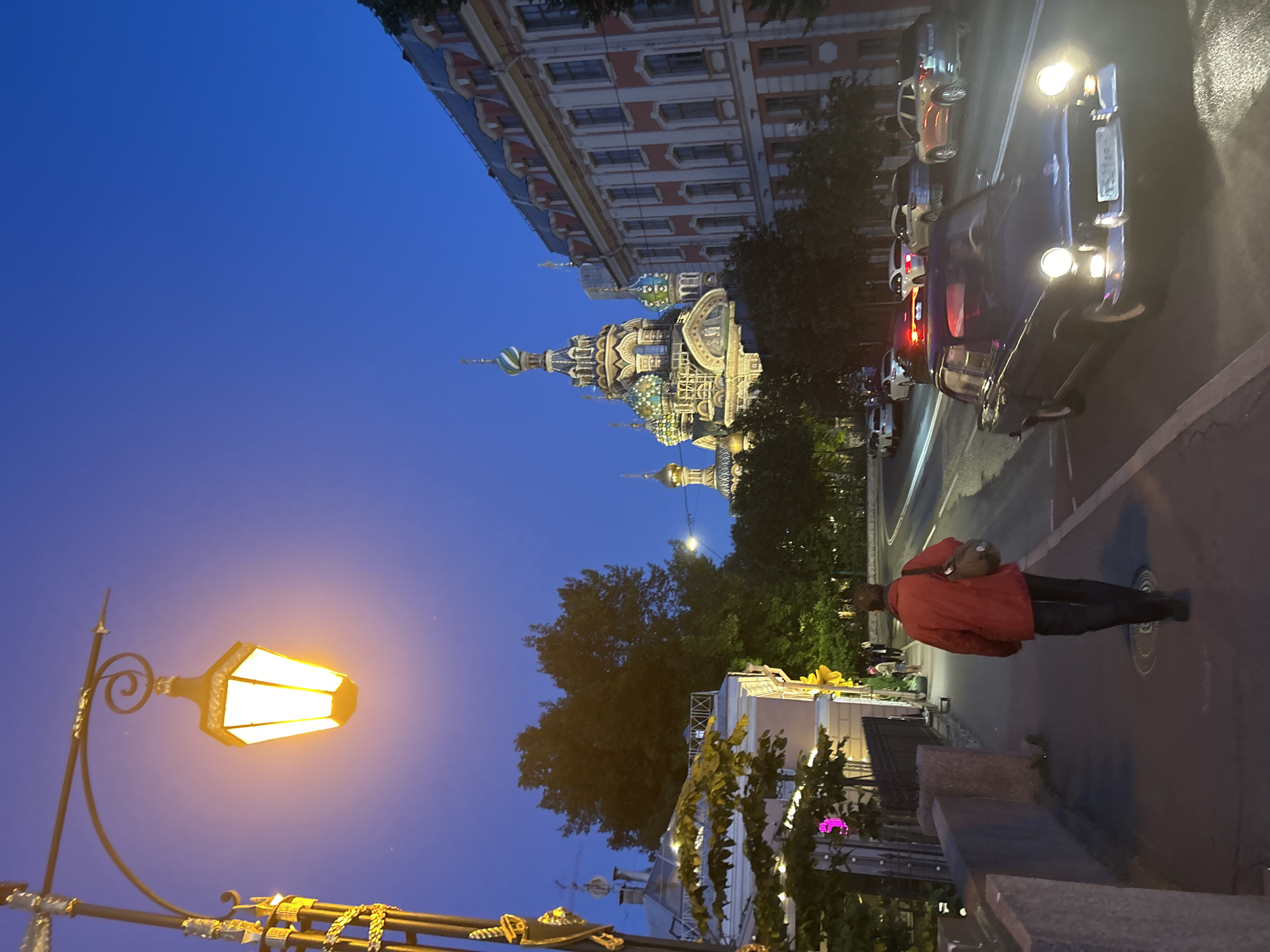An eye-level shot captures a street scene at dusk in an unknown city. A lamppost with a bright yellow light illuminates the foreground. A person wearing a red jacket and black pants walks away from the camera on a sidewalk, carrying a bag over their shoulder. The sidewalk is bordered by a low wall. Across the street, a vintage dark-colored car drives towards the viewer, its headlights on. Several other cars are visible further down the road, some with their headlights on. On the right side of the image, a grand building with red and white facades and many windows is partially visible. In the background, the distinctive onion domes of a large church, adorned with various colors, rise against the deep blue twilight sky. Trees are scattered throughout the scene, some appearing dark and silhouetted against the sky. The overall atmosphere is one of a city winding down for the night, with artificial lights beginning to dominate the fading natural light.