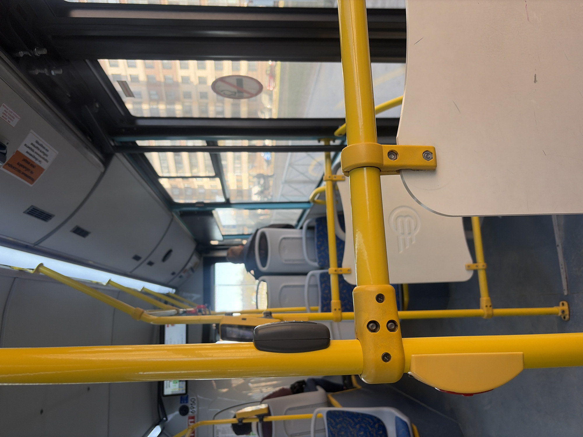 The image is taken from inside a bus in Saint Petersburg, Russia. Yellow support bars run horizontally and vertically through the frame, and a white seat back is attached to one of the vertical bars. Passengers are seated in the background, and the bus has a glass roof, through which buildings and a no-smoking symbol can be seen. The interior of the bus is clean and modern.