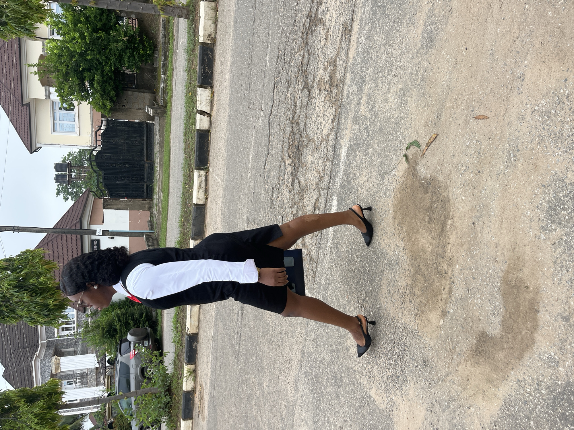 A young Black woman is walking on a paved road in Abuja, Nigeria. She is wearing a white shirt with a black vest and a black skirt. She has on glasses and is carrying a black bag. The woman is walking from left to right in the frame. In the background, there are houses and trees. The road is cracked and worn.