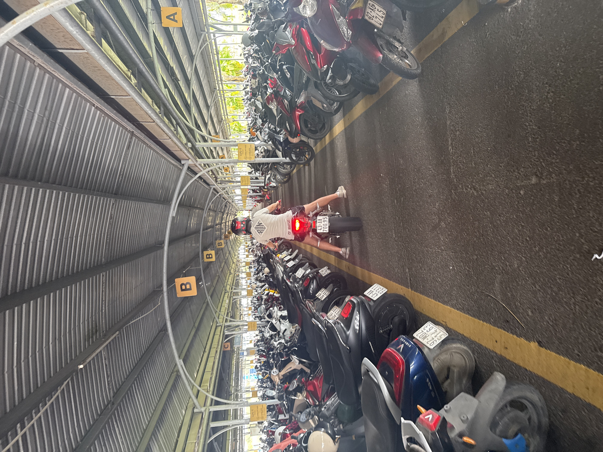 The image shows a motorcycle parking area in Đà Nẵng, Vietnam. The parking area is covered by a corrugated metal roof supported by metal arches. Numerous motorcycles are parked on both sides of a central aisle, with yellow lines marking the parking spaces. A person is riding a motorcycle down the aisle. The motorcycle has a license plate visible (43-AA 700.95), which is a registration code for Đà Nẵng. There are signs hanging from the roof, labeled "A," "B," and "C", indicating parking zones. Other visible license plates include "92-K6 4745". "92-MD2 514.63", "923.11", "92-01 055.81" and "061.86". A sign in Vietnamese "LƯU Ý" is also visible.