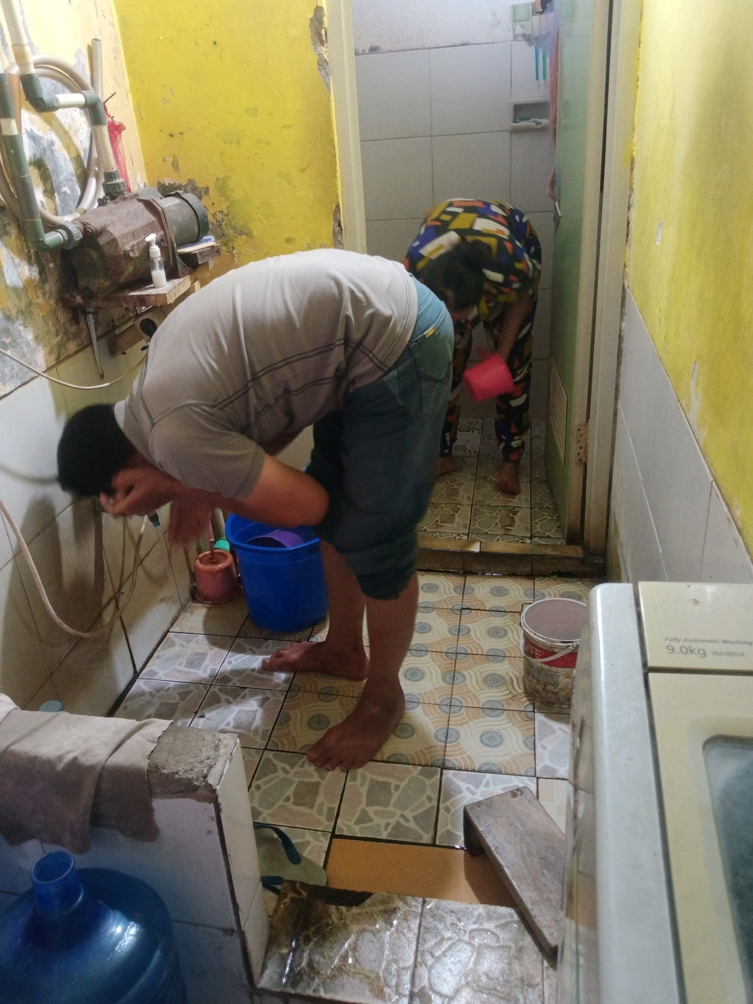 In a dimly lit, cluttered bathroom, likely in Grendeng, Indonesia, two individuals are engaged in washing activities. In the foreground, a man with dark hair, wearing a grey t-shirt and blue shorts, is bending over and washing his face. He is standing barefoot on a tiled floor adorned with a patterned design. To his left, a blue bucket with a purple ladle sits on the floor. Behind him, a rusty water pump is mounted on a yellow, deteriorating wall. In the background, a second individual, wearing a brightly colored, patterned outfit, is also bent over, appearing to be washing something, possibly with a red bucket held in their hands. The environment is characterized by worn walls, tiled flooring, and various household items scattered around, suggesting a functional but not pristine living space. A washing machine is partially visible on the right side of the frame.