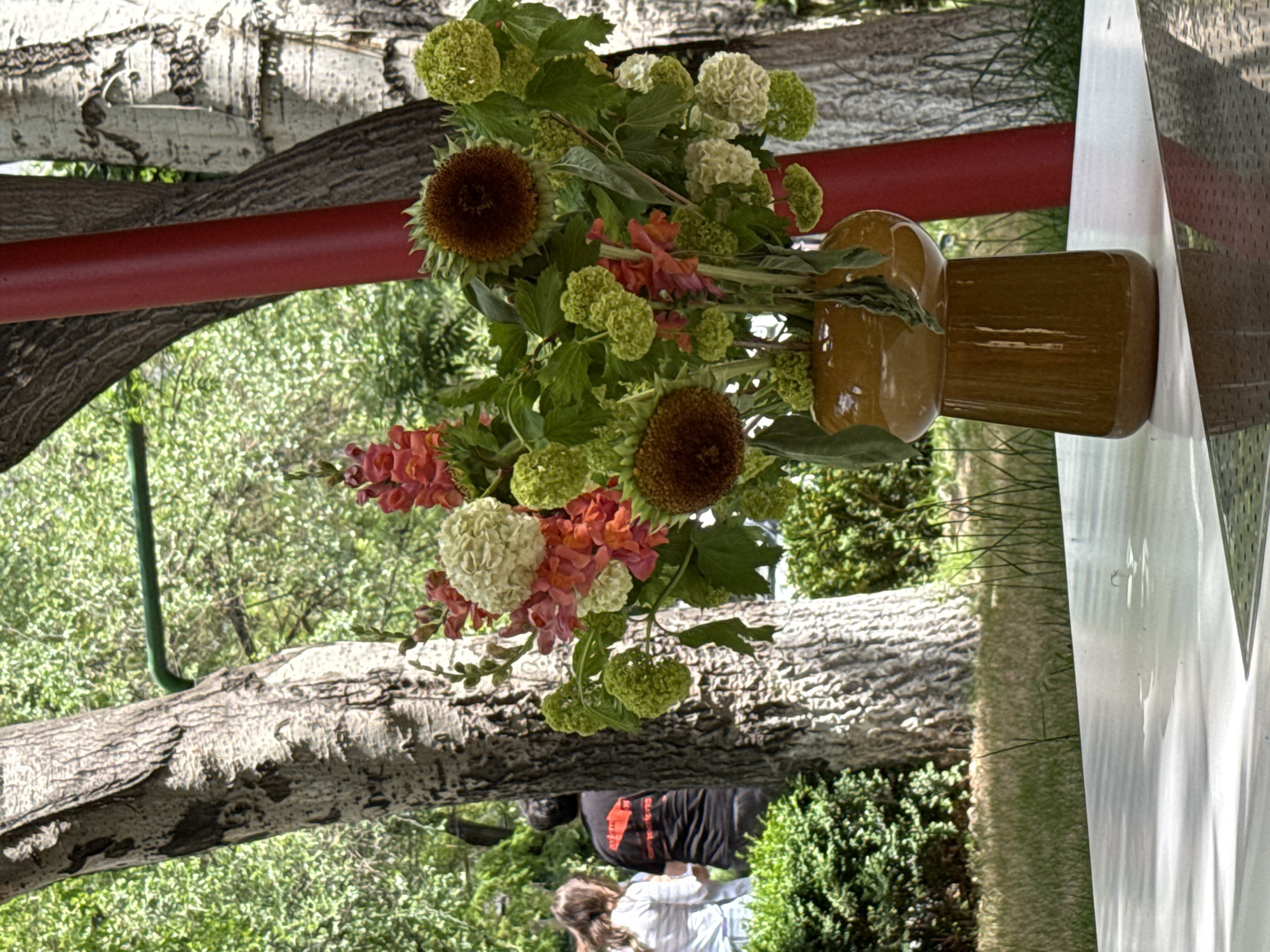 A vase of flowers, comprised of sunflowers, green hydrangeas, and pink flowers, sits on a table outdoors. People are present in the background. The setting is outdoors. The location cannot be determined.