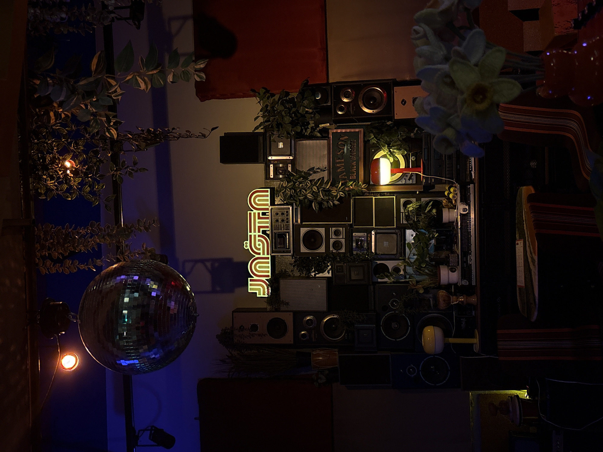 The image captures an interior scene, likely within a bar or club in Belgrade, Serbia. Dominating the left side is a disco ball hanging from the ceiling, reflecting light and creating a festive atmosphere. To the right, a wall is adorned with a collection of vintage speakers and various electronic components. A neon sign spelling "JUST" is placed vertically among the speakers. Several plants are interspersed throughout the arrangement, adding a touch of greenery. A Jameson whiskey sign can also be seen. The overall lighting is dim and moody, creating a nightclub ambience. There are no people visible in the frame.