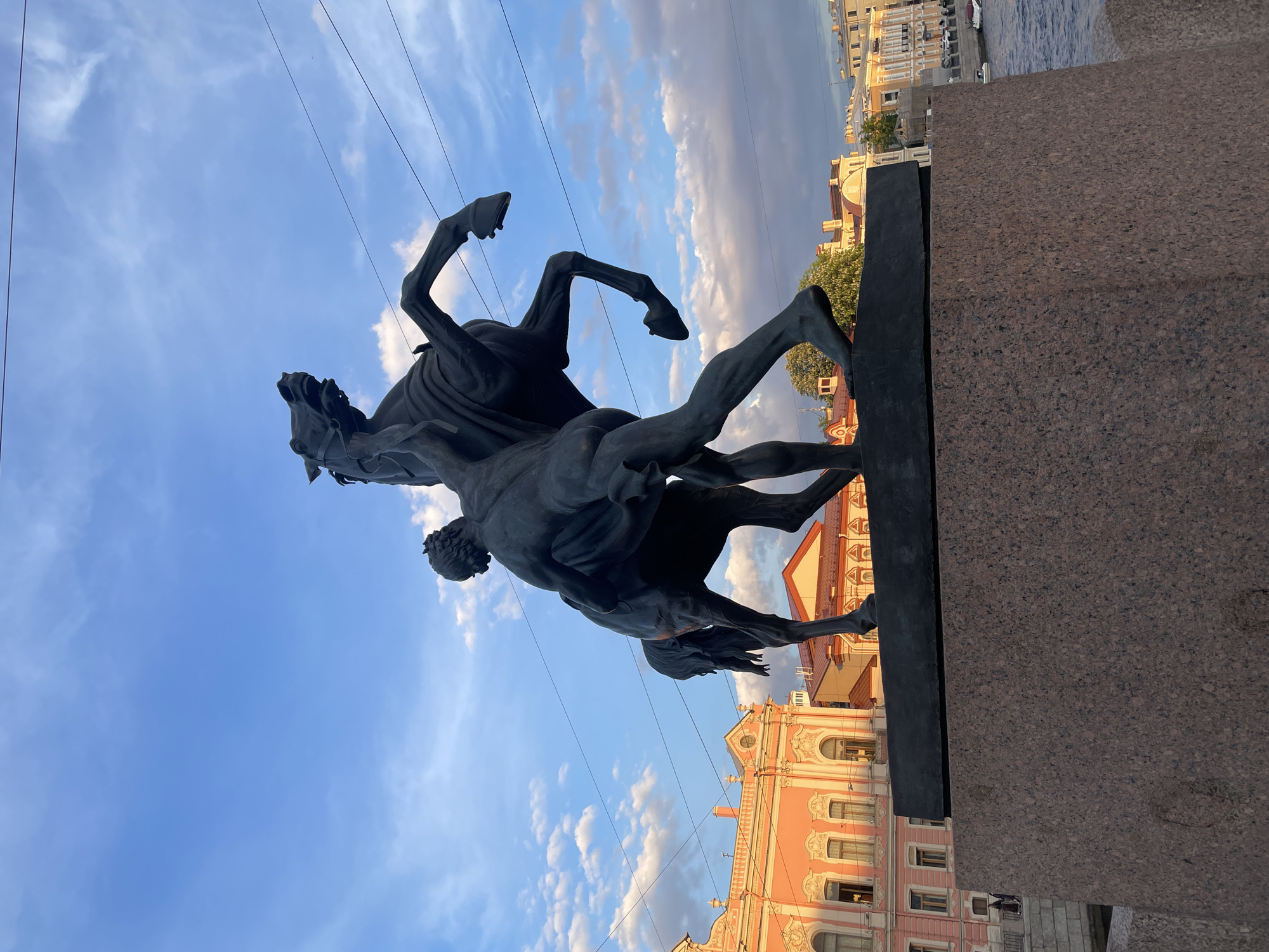A vertical, dynamic outdoor shot features a dark bronze equestrian statue against a partly cloudy blue sky, captured from a low angle. The statue depicts a powerful, rearing horse struggling with a muscular man attempting to control it. The horse is depicted mid-leap, with its front hooves lifted high, head thrown back, and mouth open. The man is positioned beneath the horse, appearing to wrestle with it, holding onto its reins or neck. The statue rests on a dark, reddish-brown granite pedestal, which occupies the lower right portion of the image.
In the background, beyond the statue and pedestal, is a cityscape with classical European architecture. Buildings with salmon-pink and yellow facades, intricate white detailing, and orange or red roofs are visible, characteristic of Saint Petersburg. A body of water, likely a canal or river, can be seen to the far right, with a small white boat docked. Overhead wires are faintly visible stretching across the sky in the upper left.
The lighting suggests late afternoon or early evening, with warm light illuminating parts of the buildings and clear blue skies with scattered cumulus clouds. The scene conveys a sense of dramatic struggle and artistic power, set within a historic urban environment. No visible text is present in the image.