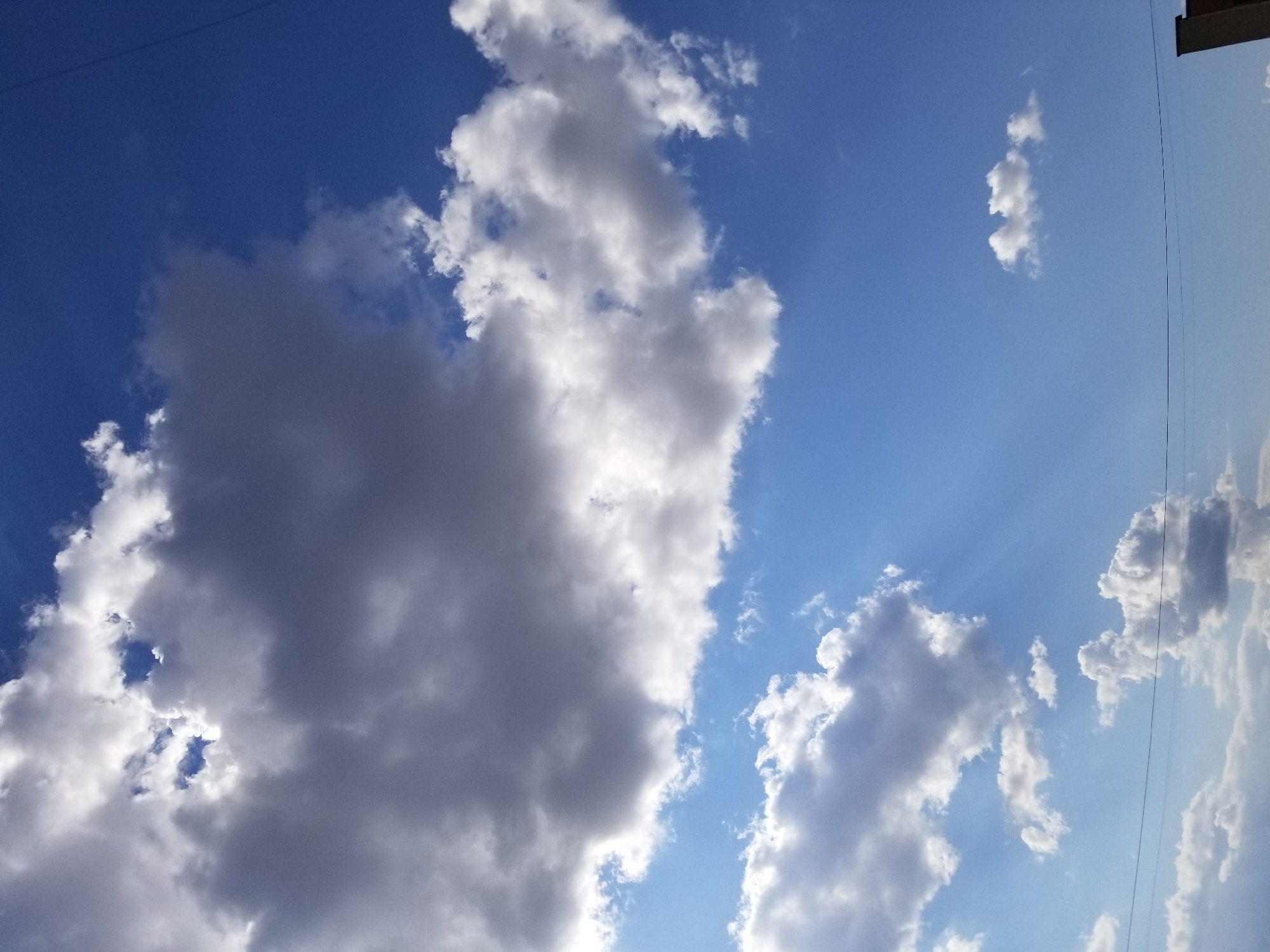 The image depicts a cloudy sky over Tiraspol, Moldova. Dominant dark gray clouds are positioned in the upper half of the frame, with patches of bright blue sky peeking through. Lighter, fluffy white clouds are scattered across the lower half. Sunlight beams are visible, creating a dramatic effect. Two horizontal power lines stretch across the lower portion of the sky, with a sliver of a building visible at the bottom right corner.