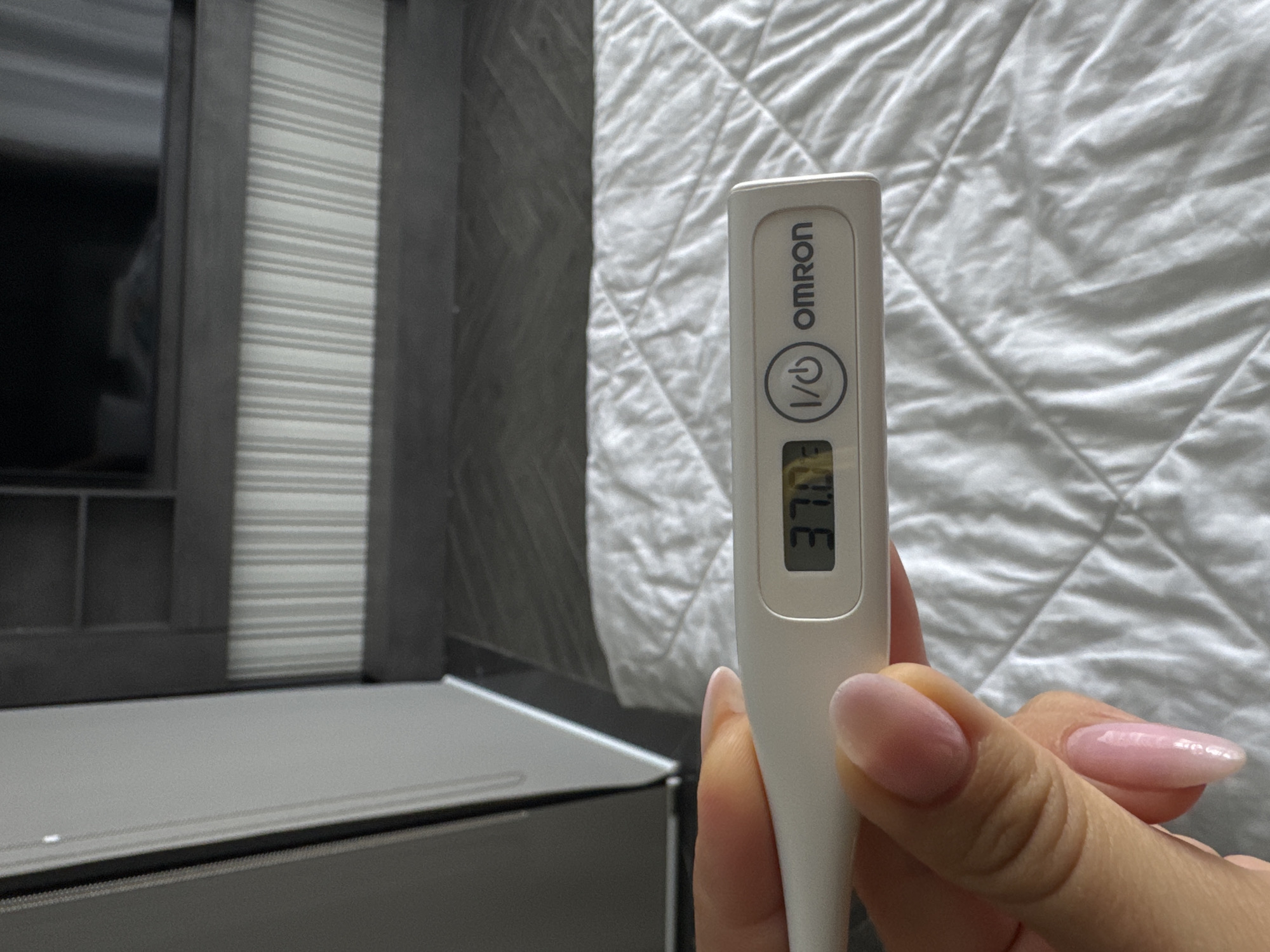 The image shows a person holding a white Omron digital thermometer displaying a temperature of 37.0 degrees Celsius. The person's hand with manicured nails is partially visible. The background includes a wrinkled white bed sheet and a gray-toned object with a striped pattern, possibly a cabinet or piece of furniture. The location is unknown.