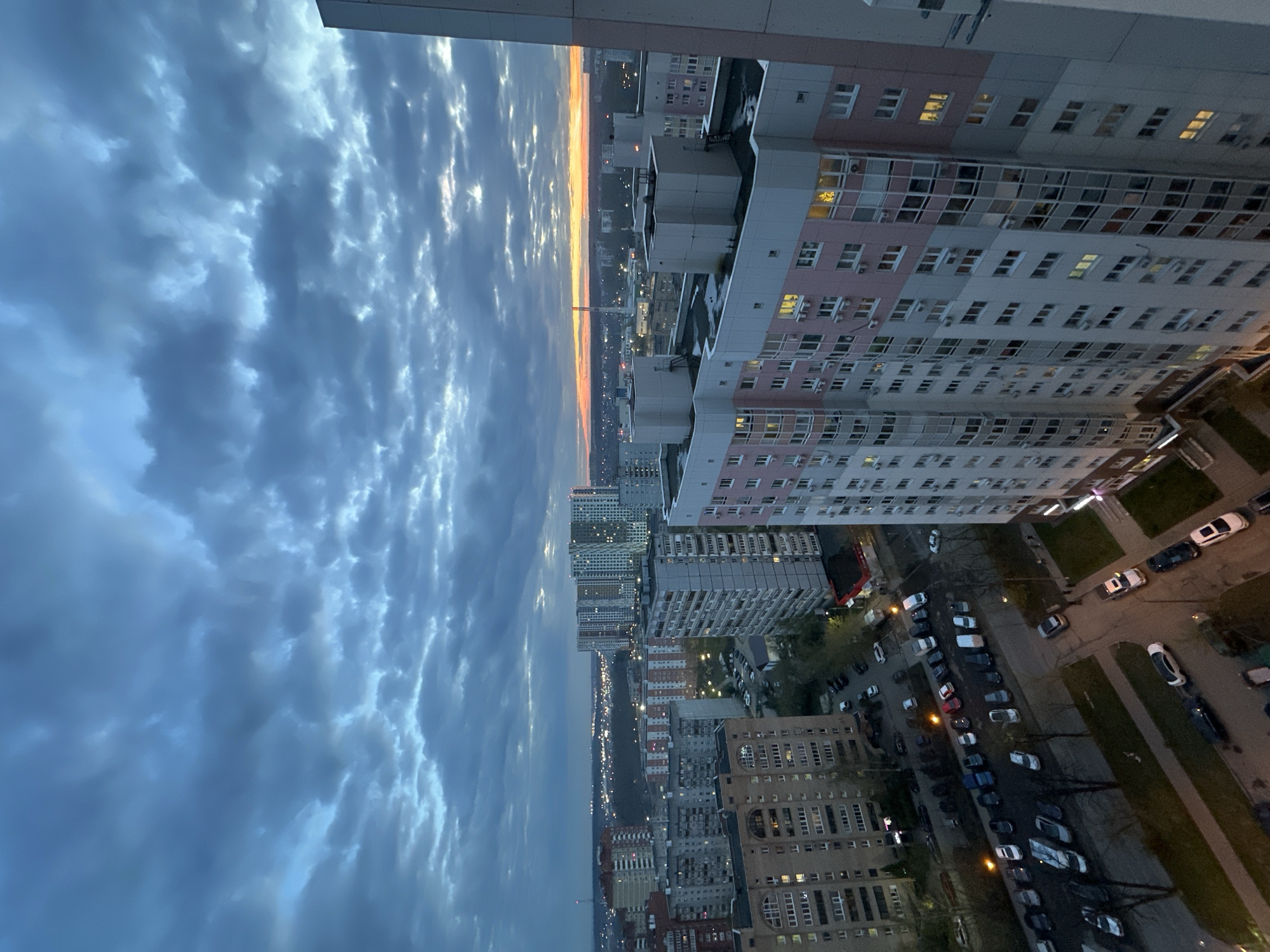 The image shows a high-angle view of a city at dusk. The foreground is dominated by a tall apartment building with pink and white facade and numerous windows. Below, streets lined with parked cars and more buildings are visible. In the distance, a skyline is faintly illuminated by the setting sun. The sky is filled with heavy, dark clouds. Based on the architecture and overall urban landscape, the location is most likely in Moscow, Russia. There are no people visible in the image. The primary subjects are the city buildings, streets, and sky. The action is a static scene of a city at dusk. There are no apparent interactions depicted. The environment is a densely populated urban area with an overcast sky.
