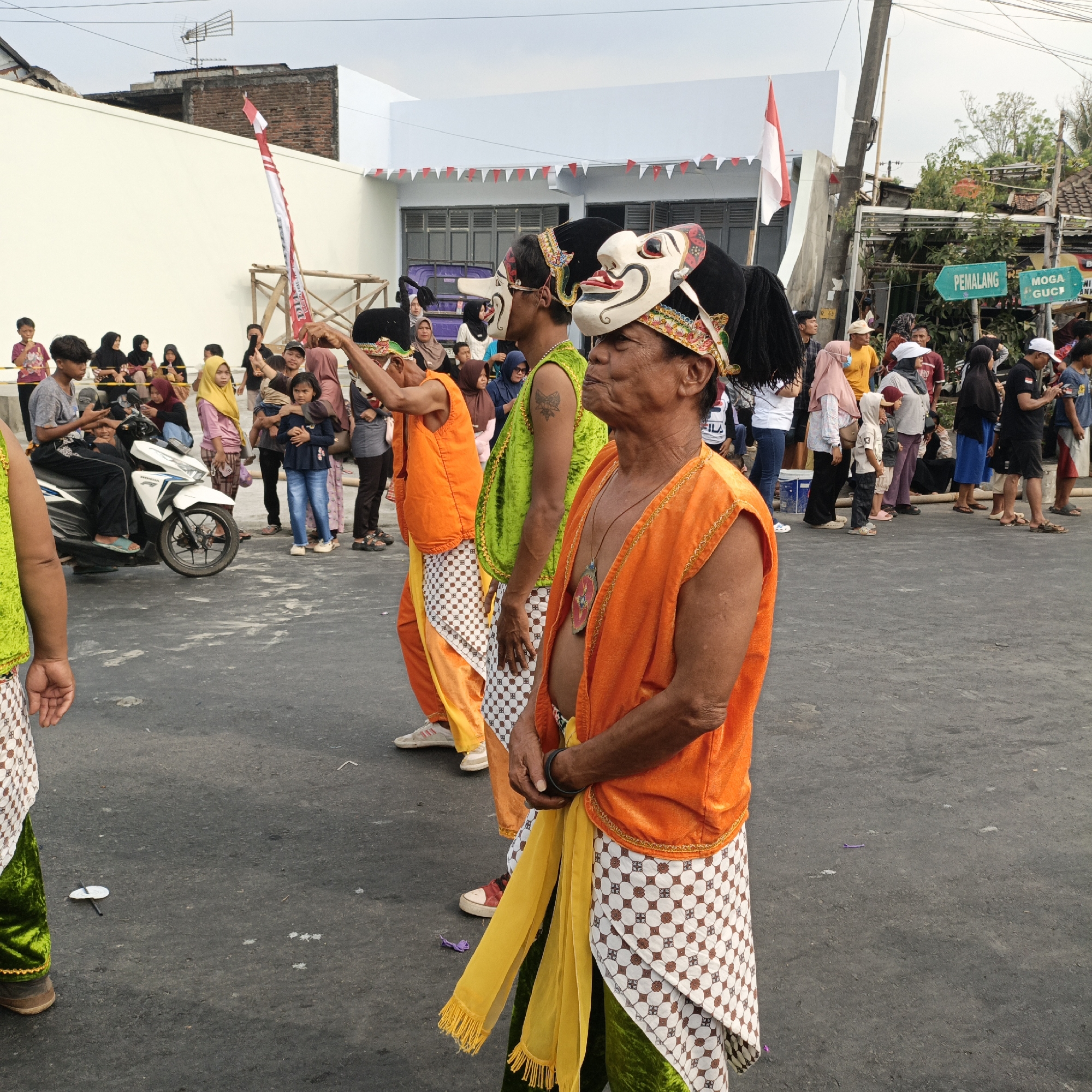The image depicts a street scene in Randudongkal, Indonesia, featuring individuals participating in a cultural performance. In the foreground, three men are dressed in traditional attire, wearing colorful masks, orange vests over green garments, and patterned skirts. The man on the right is closer to the camera and his mask has a white face with red and black accents. The man in the middle wears a green vest and a white mask with elaborate red and gold detailing. The man on the left is mostly obscured, but his orange vest and patterned skirt are visible. They are standing on an asphalt road.
In the background, a crowd of people are gathered, some standing and others seated. A white and blue scooter is parked on the left side of the road. Behind the crowd, there are buildings, including one with a white facade and red and white triangular flags strung along its roofline. A tall pole with green directional signs, one reading "PEMALANG," is also visible. A red and white Indonesian flag is prominently displayed on a pole in the upper left quadrant of the image. The sky is overcast. The overall impression is of a community event or festival taking place outdoors.