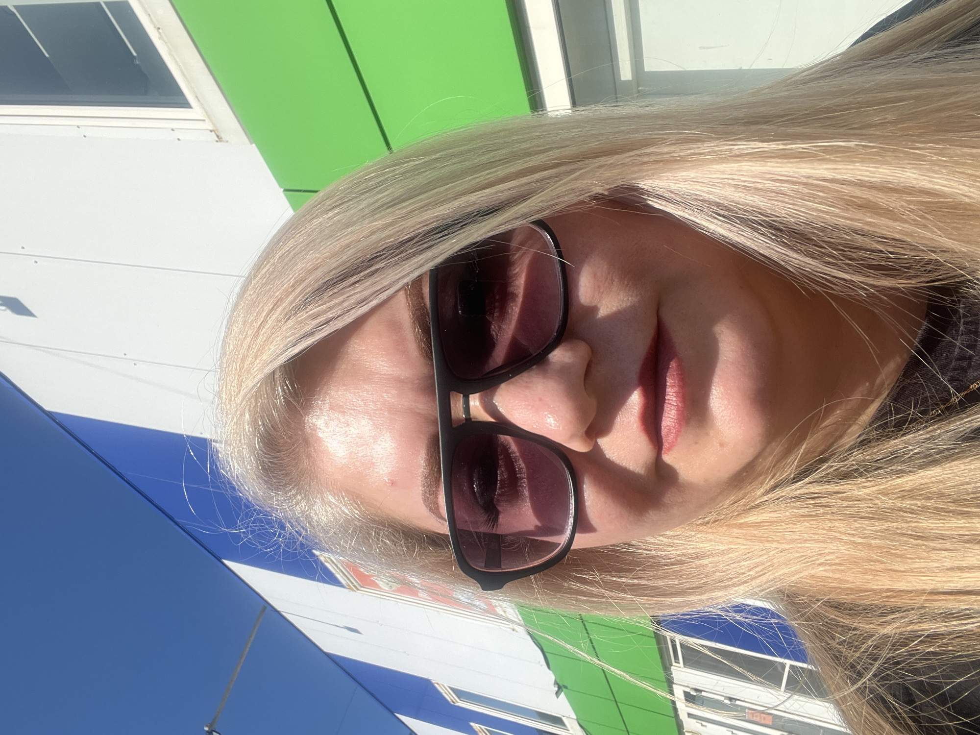 A woman with long blonde hair is wearing sunglasses and is facing the camera. The background shows a building with green, white, and blue panels. The location appears to be Glazov, Russia based on the building's design and coloring.