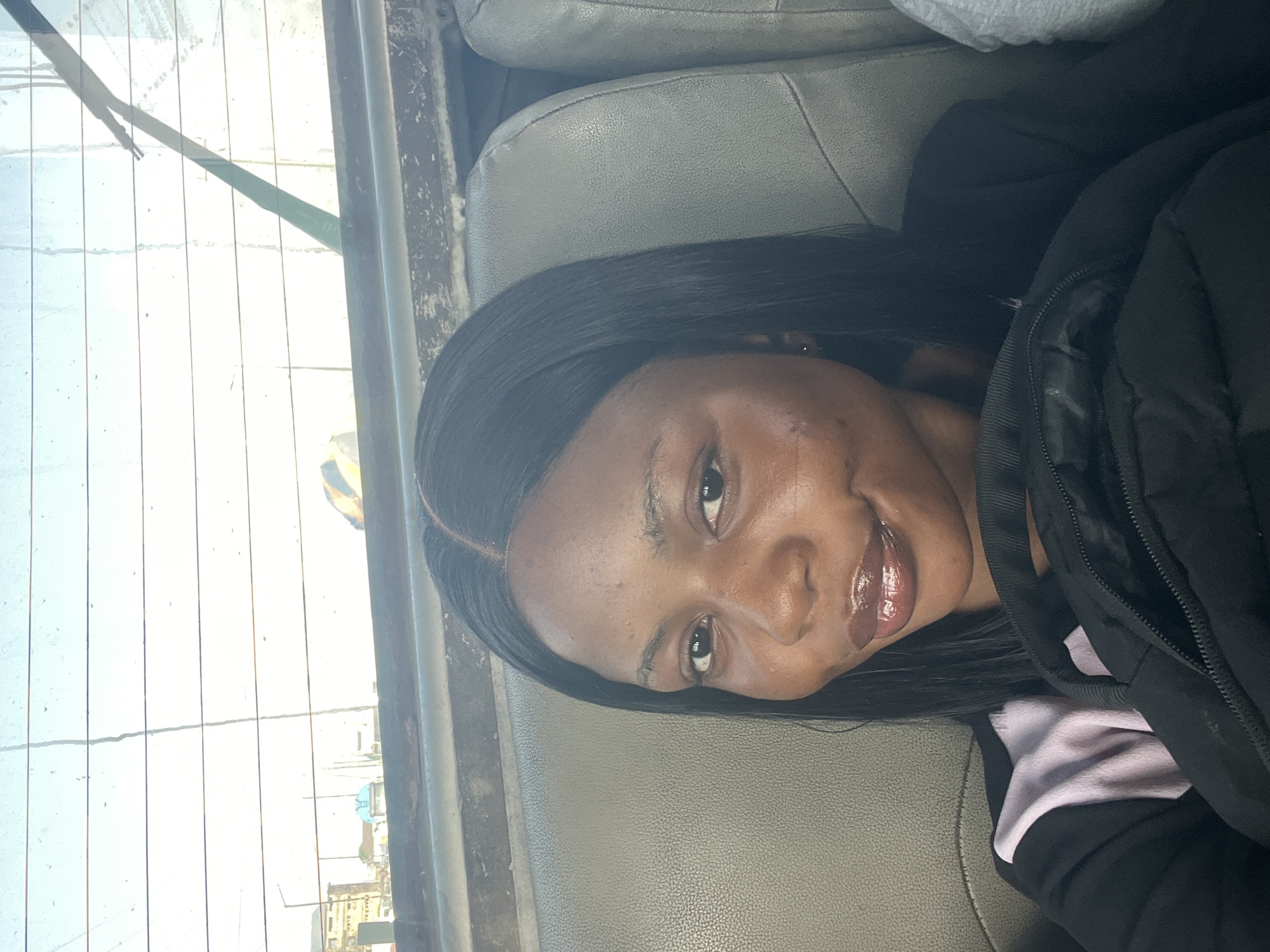 A close-up, portrait-style photograph shows a young woman seated inside a vehicle. The image is oriented sideways, with the woman looking towards the viewer and smiling with a bright, closed-mouth expression. She has dark skin, long, straight black hair with a visible part, and appears to be wearing lip gloss. She is dressed in a black jacket or coat, with a glimpse of a light purple or pink garment underneath.
The setting appears to be the interior of a car or bus, characterized by light grey or tan, textured seats visible behind and to her side. Bright daylight streams in through a window behind her, illuminating the scene. Vertical lines, likely from a rear defroster or window structure, are prominent on the glass. Through the bright, somewhat hazy window, a blurry outdoor scene suggests an urban or semi-urban environment, with indistinct shapes of buildings and possibly a street in the distance. A yellow and blue object is faintly visible outside. The strong light indicates it is daytime, likely sunny. The woman's relaxed posture and smile convey a cheerful mood. There is no visible text in the image. The location context provided is Satellite Town, Nigeria.