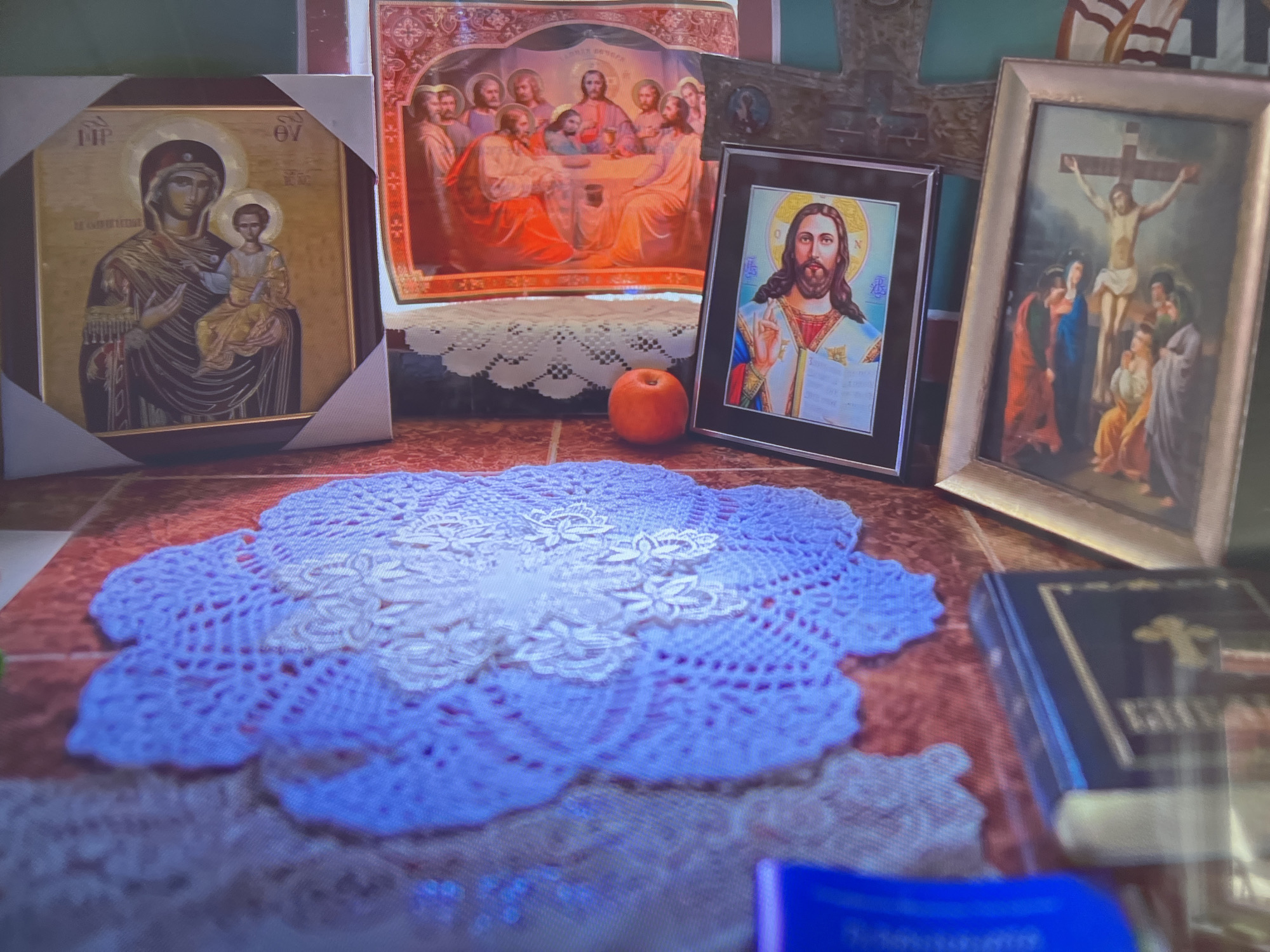 The image depicts a collection of religious icons and objects arranged on a table in an unknown location. The items include four framed images: one of the Virgin Mary holding Jesus, one of the Last Supper, one of Jesus, and one of the Crucifixion. An orange apple sits between the Last Supper image and the framed Jesus picture. A blue and white doily is prominently displayed in the foreground. A book with a golden cross on its cover is visible on the right side of the image.