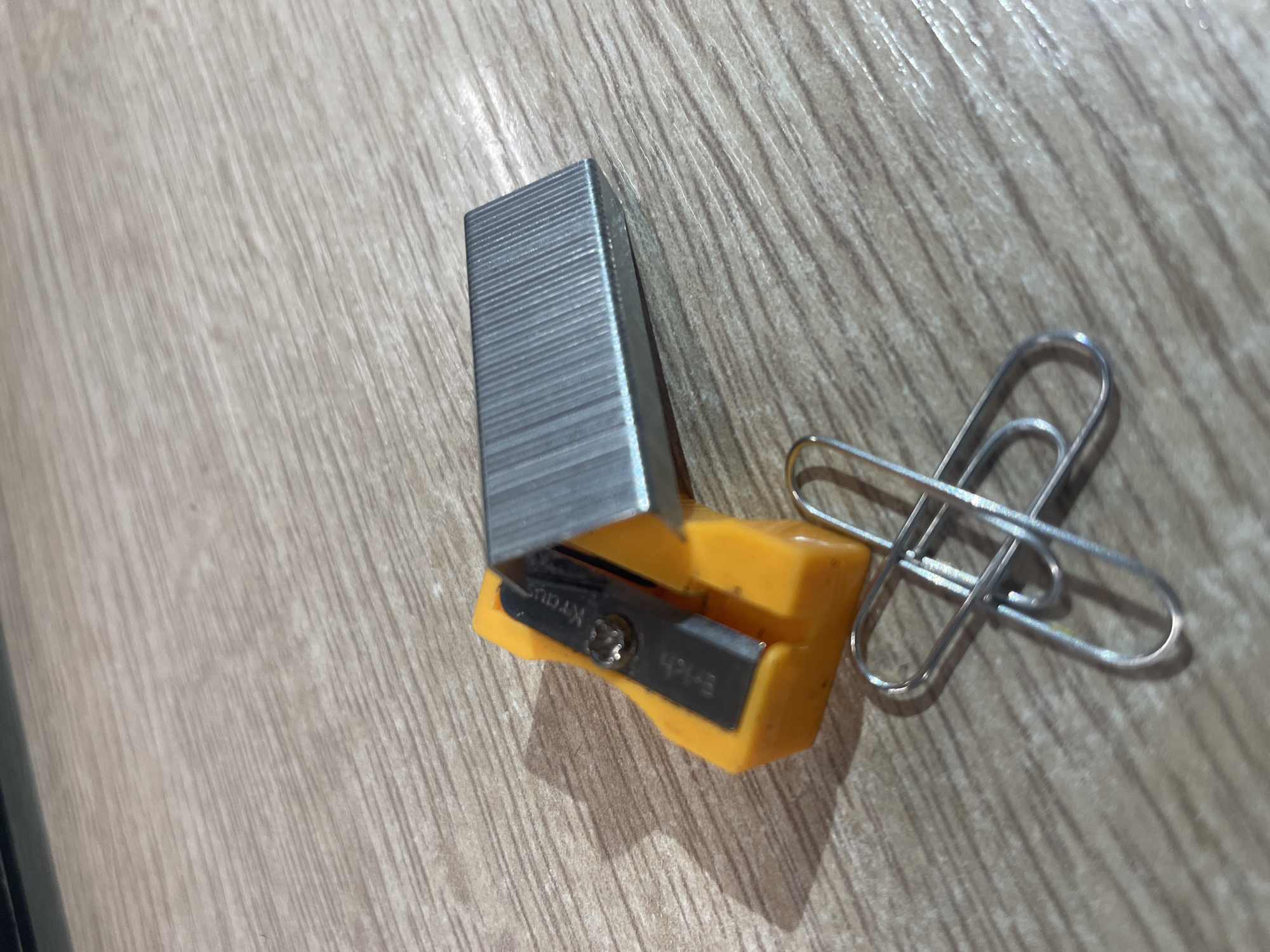 The image displays a close-up, eye-level view of office supplies on a wooden surface. The primary subjects are a yellow pencil sharpener with a metal blade, a stack of silver staples, and two silver paper clips. The pencil sharpener has the brand name "Erich Kraus" inscribed on its blade. The wooden surface appears to be a table or desk, with a visible wood grain pattern. No people or animals are present in the image, and no specific city location is identifiable from the visual information. The lighting suggests an indoor setting, possibly with artificial light.