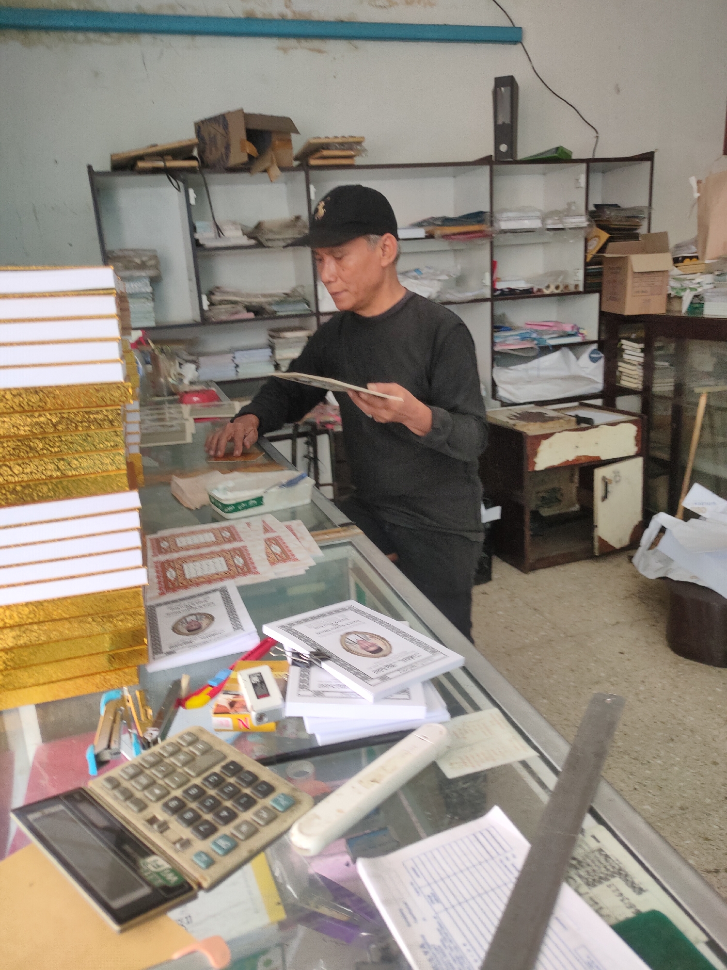 A man wearing a black baseball cap and a black long-sleeved shirt stands in a cluttered room, appearing to be a small shop or office. He is holding a thin booklet or piece of paper in his right hand and looking down at it. His left hand is resting on a counter or display case. The room is filled with shelves stacked with various items, including boxes, books, and papers. On the counter in front of him, there are more papers, a calculator, a stapler, and other office supplies. A tall stack of gold-colored books is visible to his left. A large glass display case runs across the foreground, filled with more papers and what appear to be certificates or forms. The overall impression is one of a busy, perhaps slightly disorganized, workspace. The location of Sumedang, Indonesia, is mentioned in the prompt but cannot be determined from the visual information in the image.