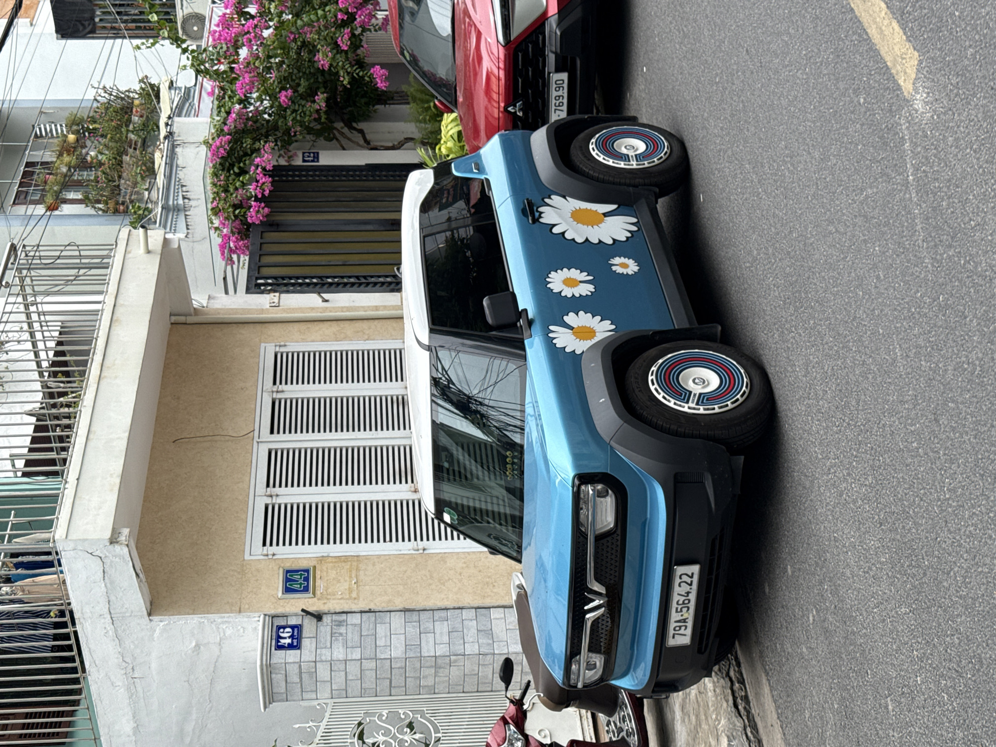 In Nha Trang, Vietnam, on a city street, a light blue electric car with white daisy decals parked next to a red SUV. The car has a white roof and black wheel wells. The license plate of the blue car reads "79A-564.22". The red SUV's license plate is partially visible, reading "-769.90". In the background, a building with a balcony and pink bougainvillea flowers is visible. To the left of the blue car, a red scooter is parked. The scene is captured from a slightly elevated perspective, showing the street, sidewalks, and surrounding buildings.