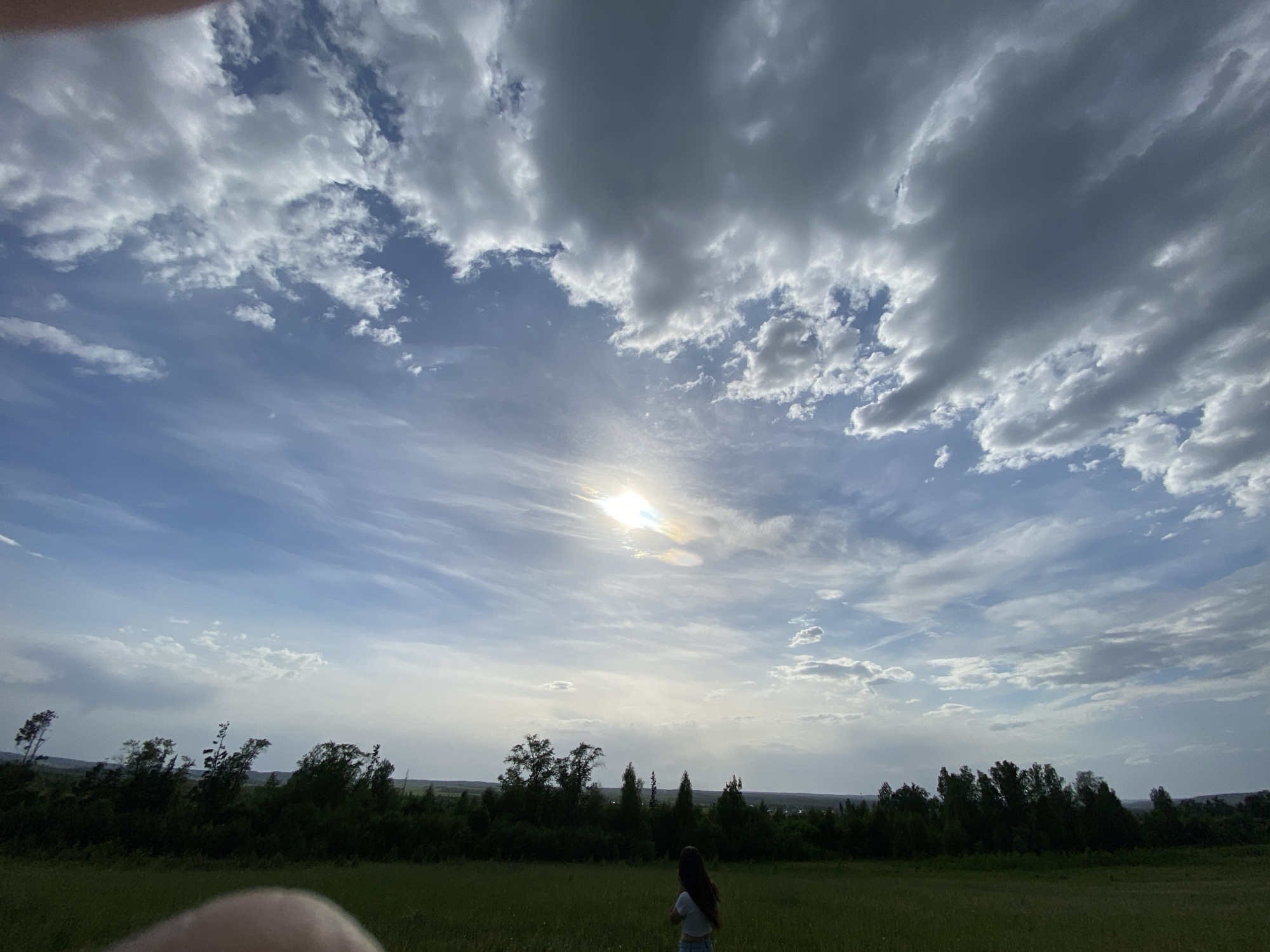 A wide-angle shot depicts a grassy field, likely in the vicinity of Гаревая, Russia, under a sky filled with dramatic clouds and the sun obscured by haze. A line of trees and shrubs marks the horizon. In the foreground, a young woman stands with her back to the camera, wearing a light-colored shirt and shorts. The lower-left corner of the image contains an out-of-focus object, possibly a finger or part of the photographer's hand.