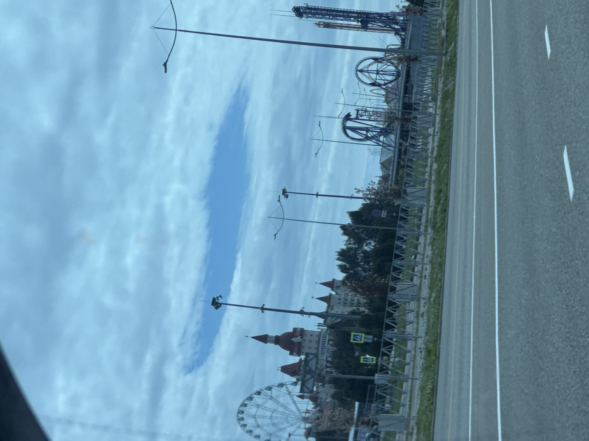 The image shows a view from a vehicle traveling on a road next to the Sochi Park amusement park in Sirius, Russia. In the background are the attractions of the amusement park, including a large Ferris wheel, a roller coaster, and a tower ride. A medieval-themed building is also visible. The sky is cloudy. The road is divided by white lines. The scene appears to be taken during the daytime.