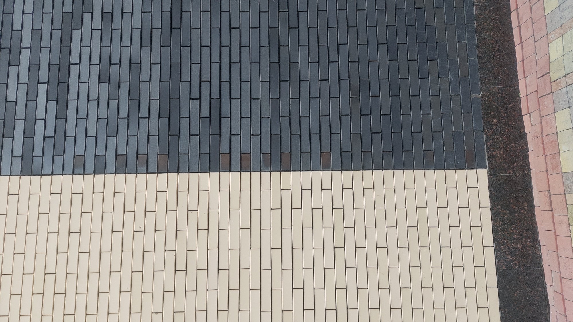 The image shows a ground surface in Novosibirsk, Russia, paved with small rectangular bricks arranged in a running bond pattern. The bricks are laid in two distinct color blocks: the upper portion is dark gray, while the lower portion is cream-colored. A dark brown, granular-textured strip runs vertically along the right edge of the image, suggesting a different paving material or a border. Adjacent to this strip are other bricks in shades of yellow, peach, and red, also arranged in a running bond pattern. The bricks appear to be set in mortar, creating distinct lines between them. No people or other objects are visible in the frame. The environment suggests a pedestrian area, possibly a sidewalk or a public square.
