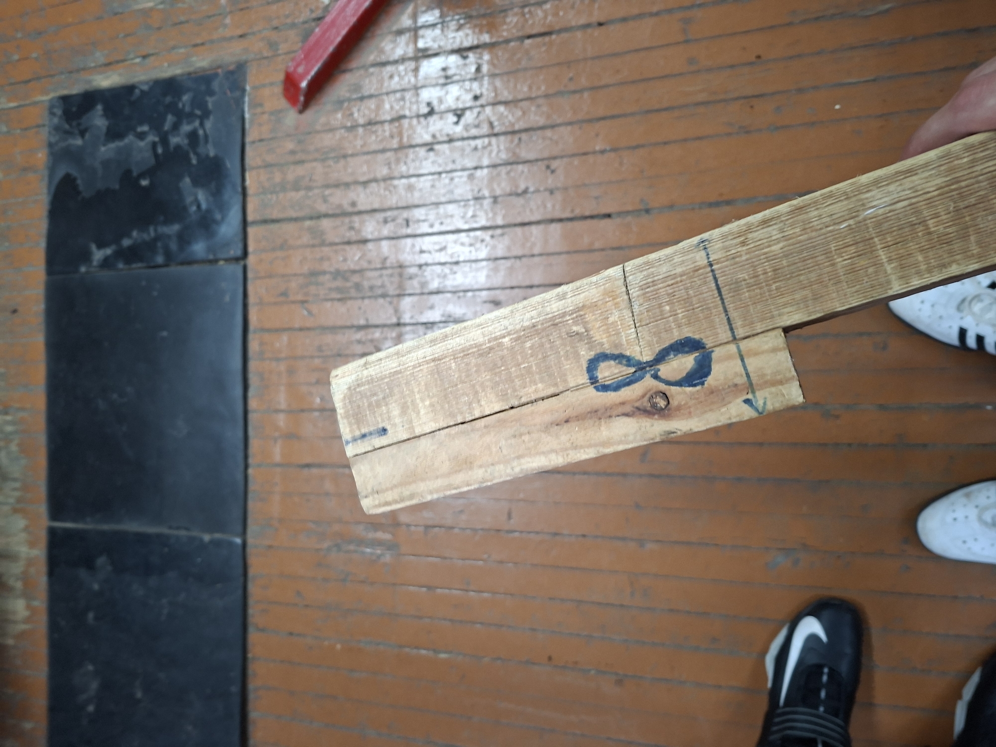 The image depicts a close-up view of a piece of wood with the number "8" drawn on it in blue marker. The wood is positioned on a wooden floor with distinct planking and a worn, orange-brown finish. In the background, there are several dark, rectangular objects, possibly tiles or mats, laid out on the floor. To the right of the wood, a red object, which appears to be a tool or a piece of plastic, is visible. On the left side of the image, a person's feet are visible, wearing a black and white athletic shoe. Further down, another person's feet are visible wearing white athletic shoes. The overall scene is indoors, with the lighting suggesting it is daytime. There is no specific information within the image to determine its location in Kazan, Russia.