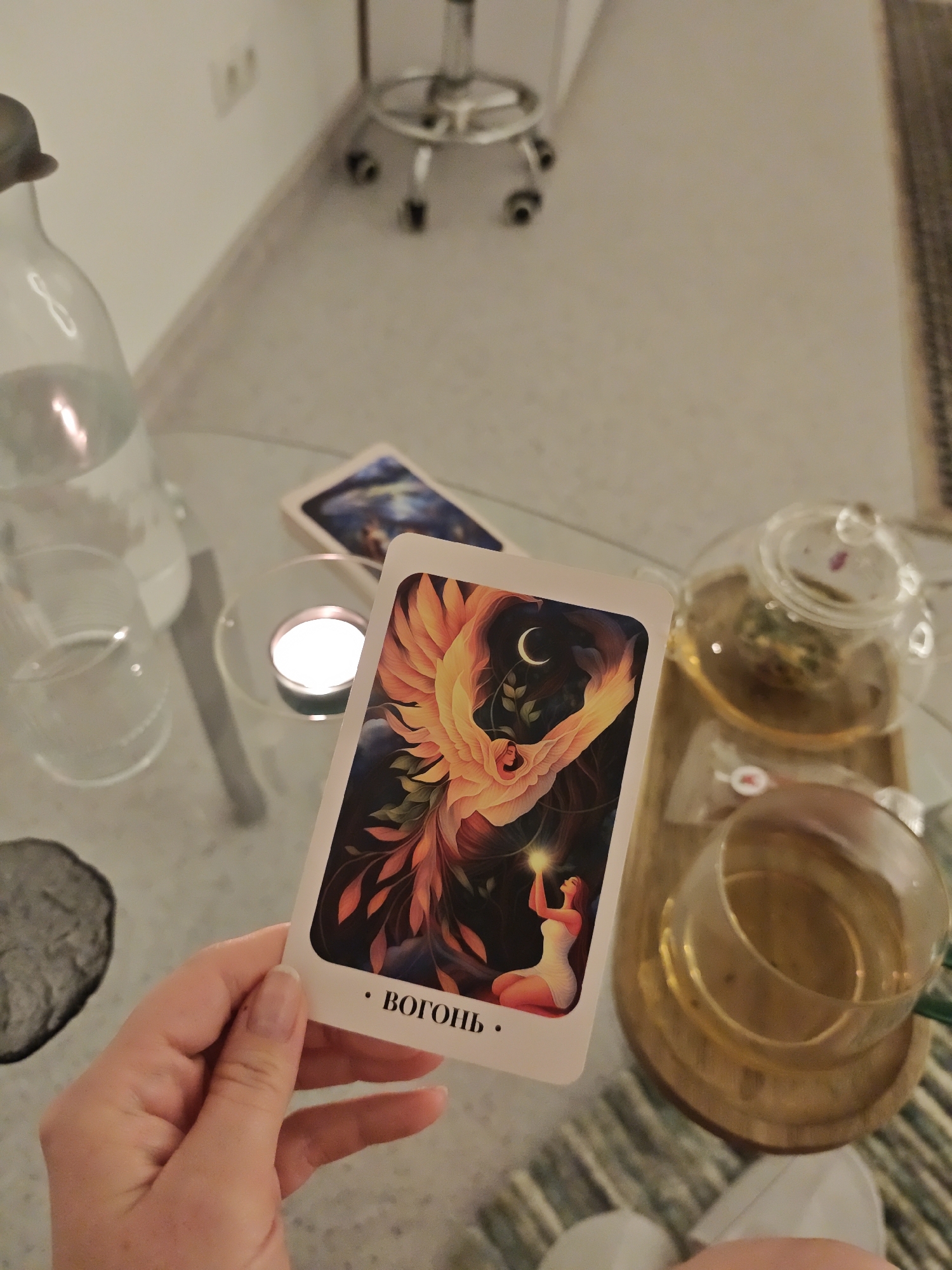 A hand holds a tarot card with an illustration of a phoenix above a kneeling woman, the word "ВОГОНЬ" (Fire) printed below. In the background, a glass table holds a bottle of water, a lit tealight candle, another tarot card, a teapot, and a cup of tea. The scene appears to be indoors. Location: Vinnytsia, Ukraine cannot be verified.