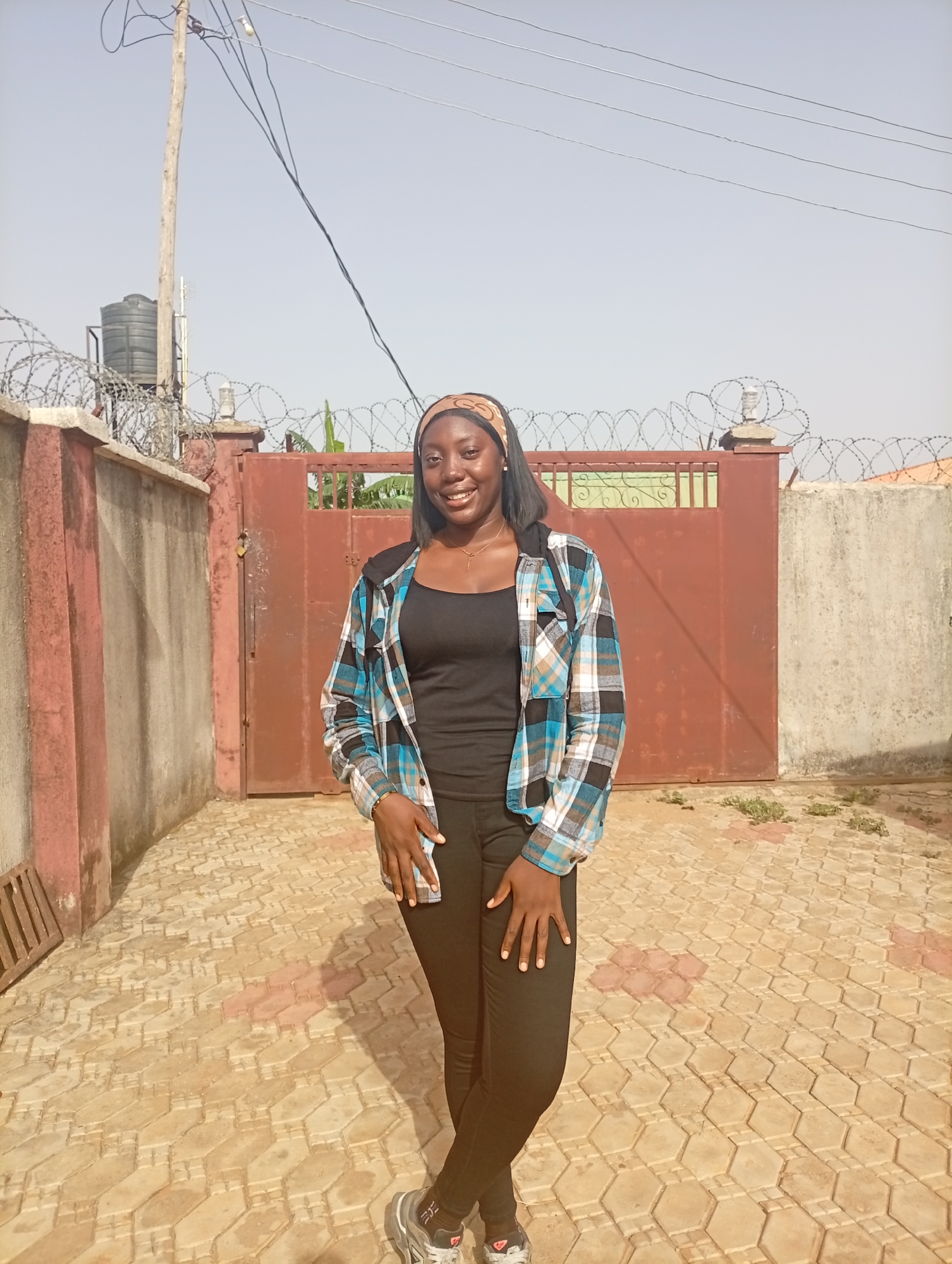 A young woman stands in a paved courtyard in Rido, Nigeria. She wears a black tank top, black jeans, a blue and white plaid shirt, and a patterned headband. She is smiling and has her hands resting on her thighs.
Behind her is a reddish-brown metal gate topped with barbed wire, set between concrete walls. A water tank is visible atop a pole behind the left wall. Power lines run above the walls and gate. The paving stones of the courtyard are primarily light brown with reddish-brown accents. The sky is a light, hazy blue.