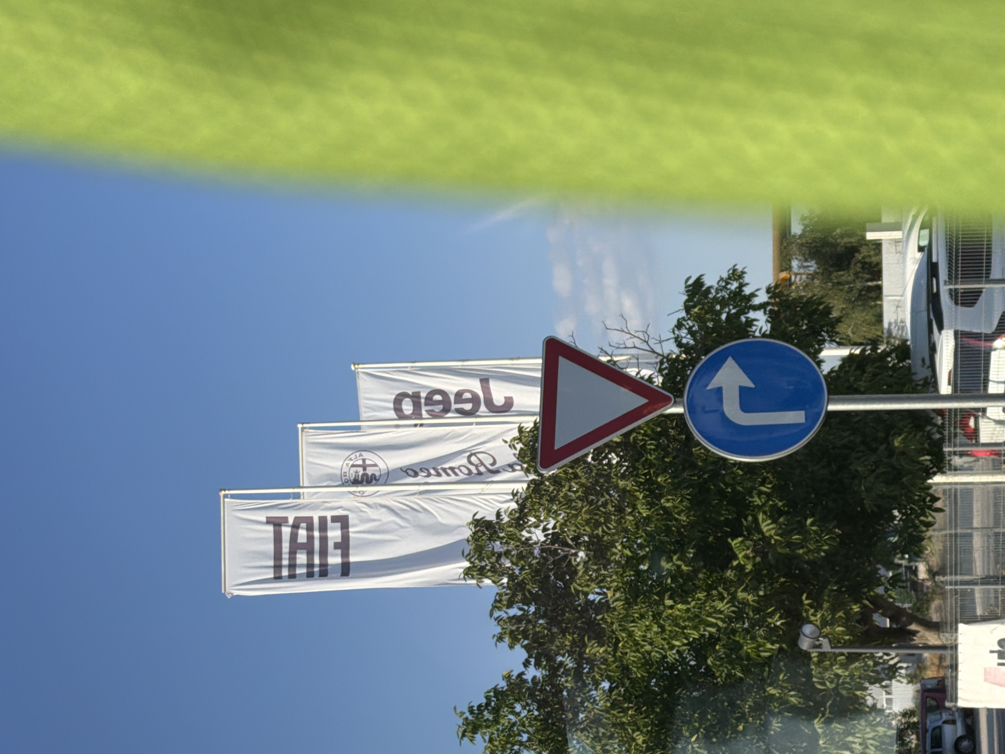 The image captures a roadside scene in Plovdiv, Bulgaria, on a clear day. The foreground is dominated by a bright green, out-of-focus element, possibly fabric or foliage, obscuring the right side of the frame. Visible in the center are two road signs: a triangular "give way" sign with a red border and a blue circular sign with a white arrow indicating a right turn. A metallic pole supports these signs. Behind the signs, a leafy green tree stands tall, its branches reaching towards the sky.
In the background, three white flags with dark lettering are prominently displayed against a bright blue sky. The leftmost flag bears the word "FIAT" in large, bold letters. To its right, another flag displays the Alfa Romeo logo and text, followed by a third flag with "JEEP" written vertically. These flags suggest the presence of a car dealership or an automotive-related business. Further in the background, a chain-link fence encloses an area where several vehicles, including a silver SUV and a white van, are parked. The overall environment appears to be an urban or semi-urban setting.