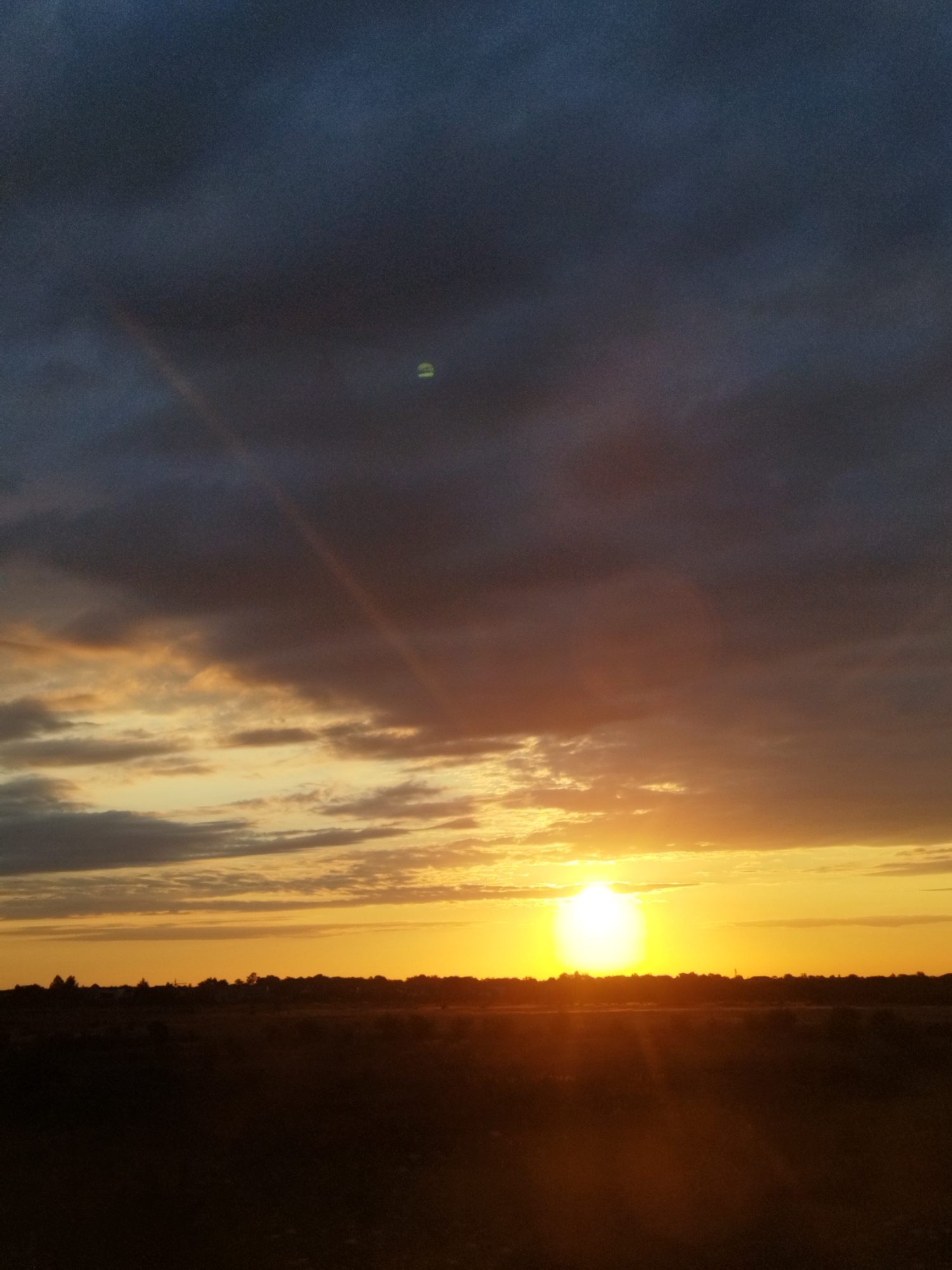 This image depicts a sunset over a rural landscape, likely in or near Chitila, Romania. The sky is filled with dramatic clouds, ranging in color from dark grey at the top to vibrant oranges and yellows near the horizon. A bright sun is visible, partially obscured by clouds, casting a warm glow across the scene. A faint rainbow-like arc of light is visible in the upper left portion of the sky. Below the sky, a dark silhouette of trees and foliage forms a horizon line. The foreground consists of a dimly lit, dark field, suggesting it is either late evening or the area is unlit. There are no people or discernible man-made structures in the immediate foreground or visible in the silhouette.