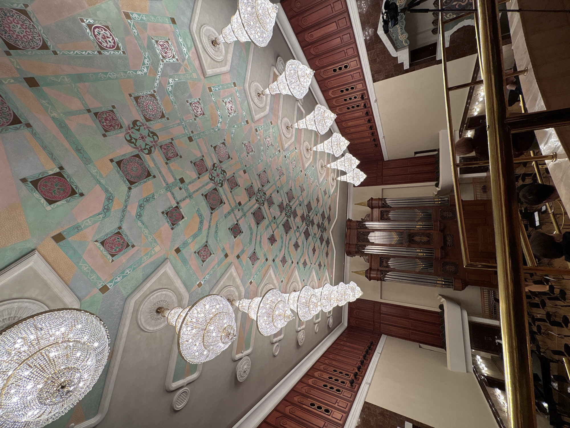 The image shows the interior of the Qolşärif Mosque in Kazan, Russia. The vantage point is from within the mosque, angled upwards towards the ornate ceiling, which is adorned with geometric patterns in muted colors, primarily pastel greens, pinks, and grays. A line of large, crystal chandeliers hangs below the ceiling. To the right of the image, a large pipe organ is visible, constructed of dark wood. A gilded railing runs partially across the right side of the frame, and glimpses of people can be seen behind it, sitting in rows. The architectural style suggests a blend of traditional Islamic and modern design elements.