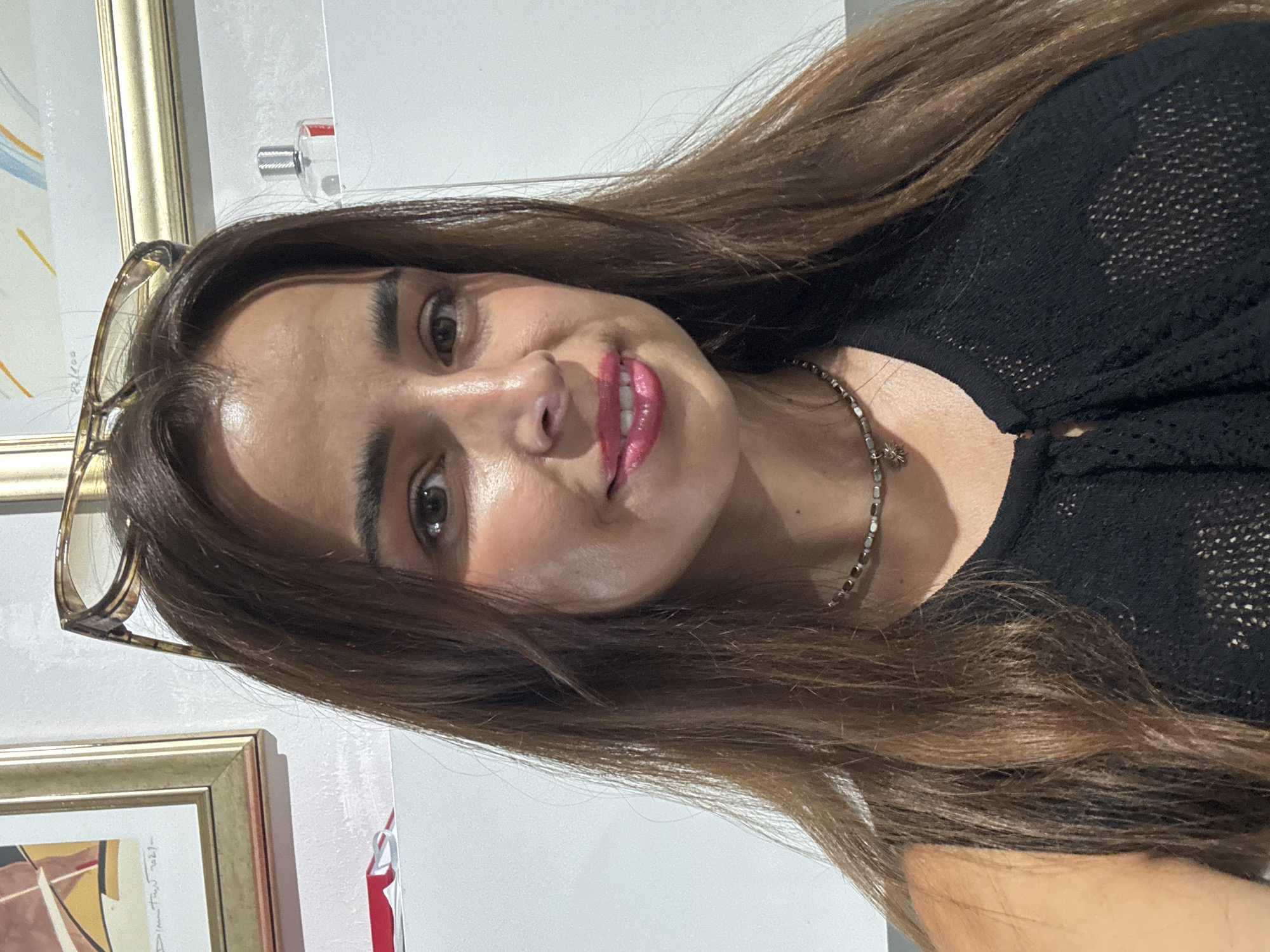 A young woman with long, brown hair and glasses perched on her head looks directly at the camera. She is wearing a black, sleeveless top with a textured pattern. Her makeup includes pink lipstick and eyeshadow. A delicate necklace with a star pendant is visible around her neck. In the background, a framed artwork and a small bottle are on a white shelf. The overall setting appears to be indoors, with a clean, modern aesthetic. The lighting suggests it might be daytime, with a light source illuminating the woman's face from the front.