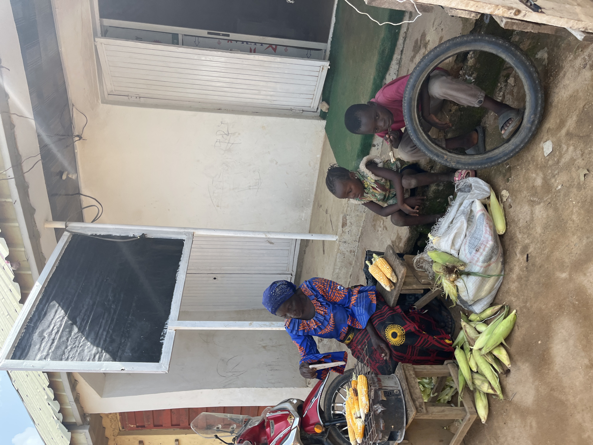 This image captures a scene in Jos, Nigeria, featuring a group of people and a marketplace stall. At the center, a woman in a vibrant blue and orange patterned dress and headwrap is tending to corn. She is holding a stick, possibly for roasting the corn, and several ears of corn are visible on a grill attached to a motorcycle. Nearby, a pile of fresh corn husks lies on the ground.
To the left of the woman, a silver metal structure with black material is visible, possibly a display or shelter for the stall. The ground is made of packed dirt and concrete.
In the background, two young children are crouched down. One, wearing a pink shirt, looks towards the camera, while the other, in a patterned garment, is looking down. A bicycle tire is prominently displayed, perhaps as a play item or part of the stall's setup. The buildings in the background appear to be simple structures with white walls and doors. The overall lighting suggests it is daytime, with bright sunlight casting shadows. The scene appears to be an outdoor market, characterized by the sale of agricultural produce and the informal setting. There is no visible text in the image.