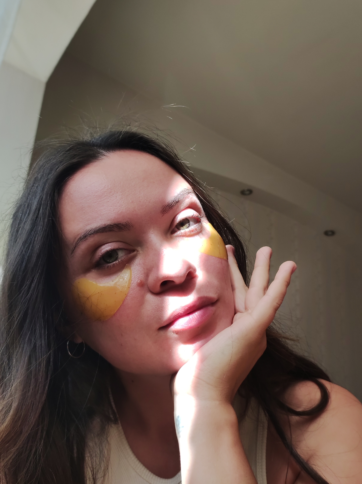 A young woman with long, dark hair is pictured from the shoulders up. She has yellow under-eye patches and is touching her face with her right hand. Sunlight is casting a shadow across her face. The background appears to be an indoor setting with neutral-colored walls and ceiling. The location is not specified.