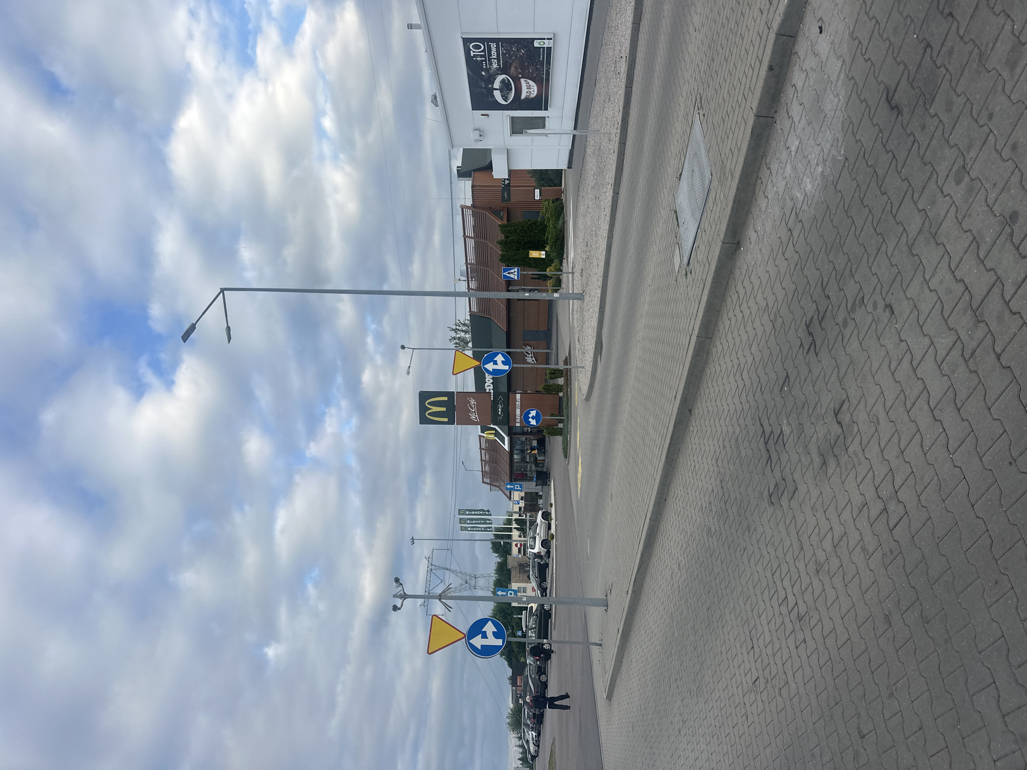 This image captures a view of a McDonald's restaurant located in Olempin, Poland. The scene is an outdoor area with a paved parking lot and sidewalk. In the background, the McDonald's building is visible, featuring its characteristic architecture and signage. A McDonald's drive-thru lane is also discernible, with cars present in the vicinity. Several traffic signs, including directional arrows and warning signs, are placed along the roadside. A street light and utility poles are also present. The sky above is partly cloudy, suggesting daytime. The ground is paved with interlocking concrete blocks. The overall scene depicts a typical fast-food restaurant environment in a commercial or roadside setting.